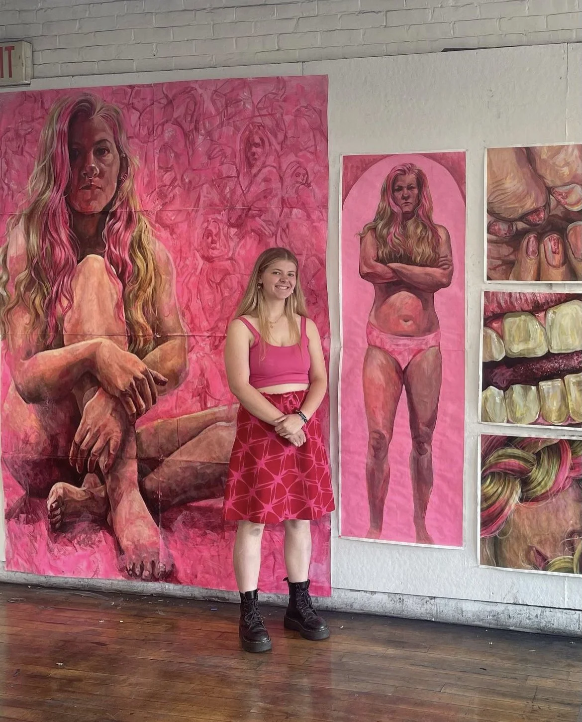 A young woman with long blonde hair, wearing a pink crop top, a pink patterned skirt, and black combat boots, standing inside an art gallery in front of pink-themed large paintings featuring female portraits and abstract art.