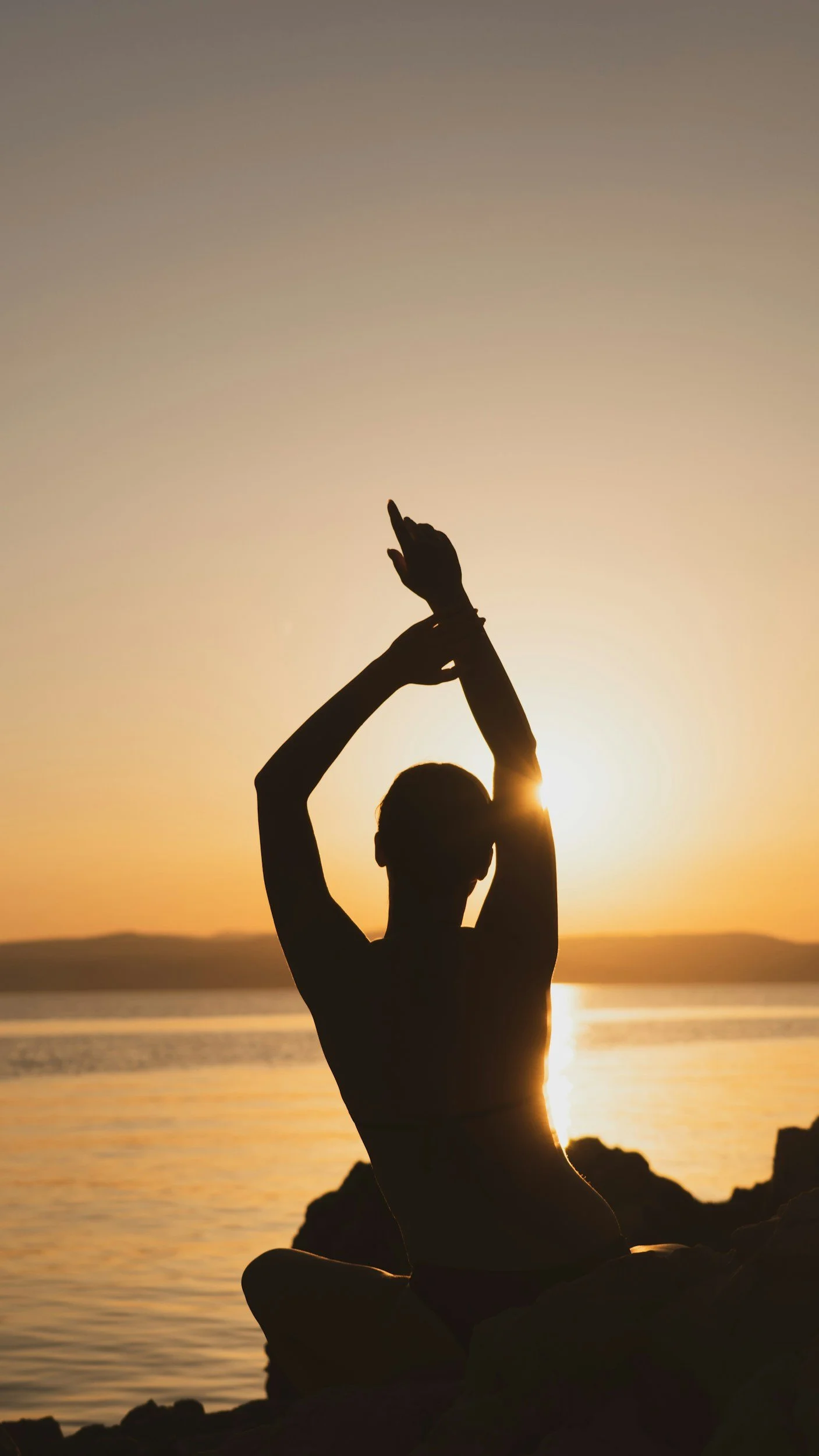 Silhouette of a woman practicing yoga near the water at sunset with the sun shining behind her.
