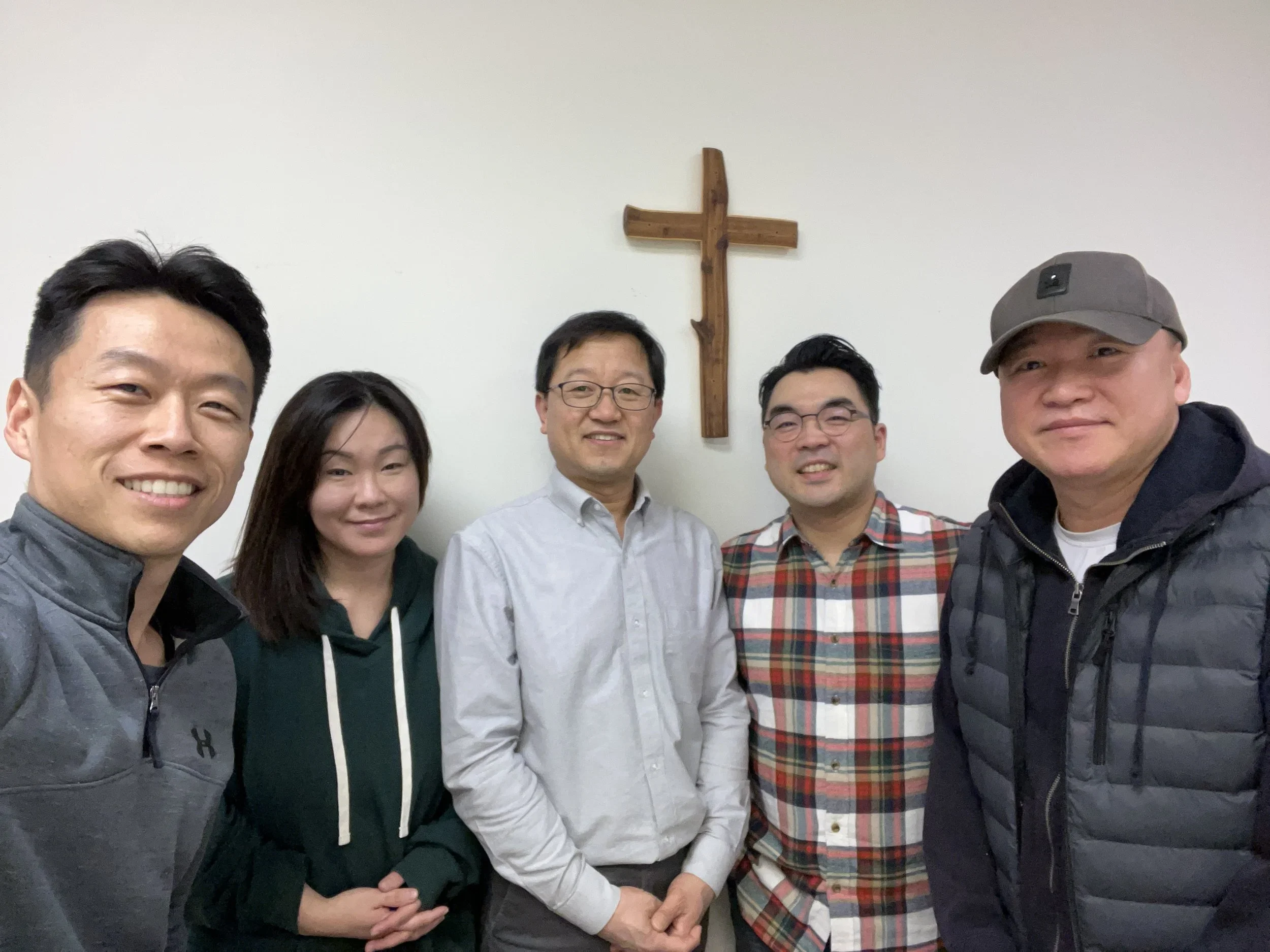 Five Korean individuals standing in front of a white wall with a wooden cross on it