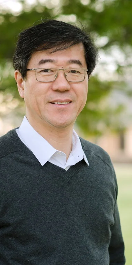 Portrait of a man wearing glasses, a white collared shirt, and a dark sweater, standing outdoors with trees in the background.