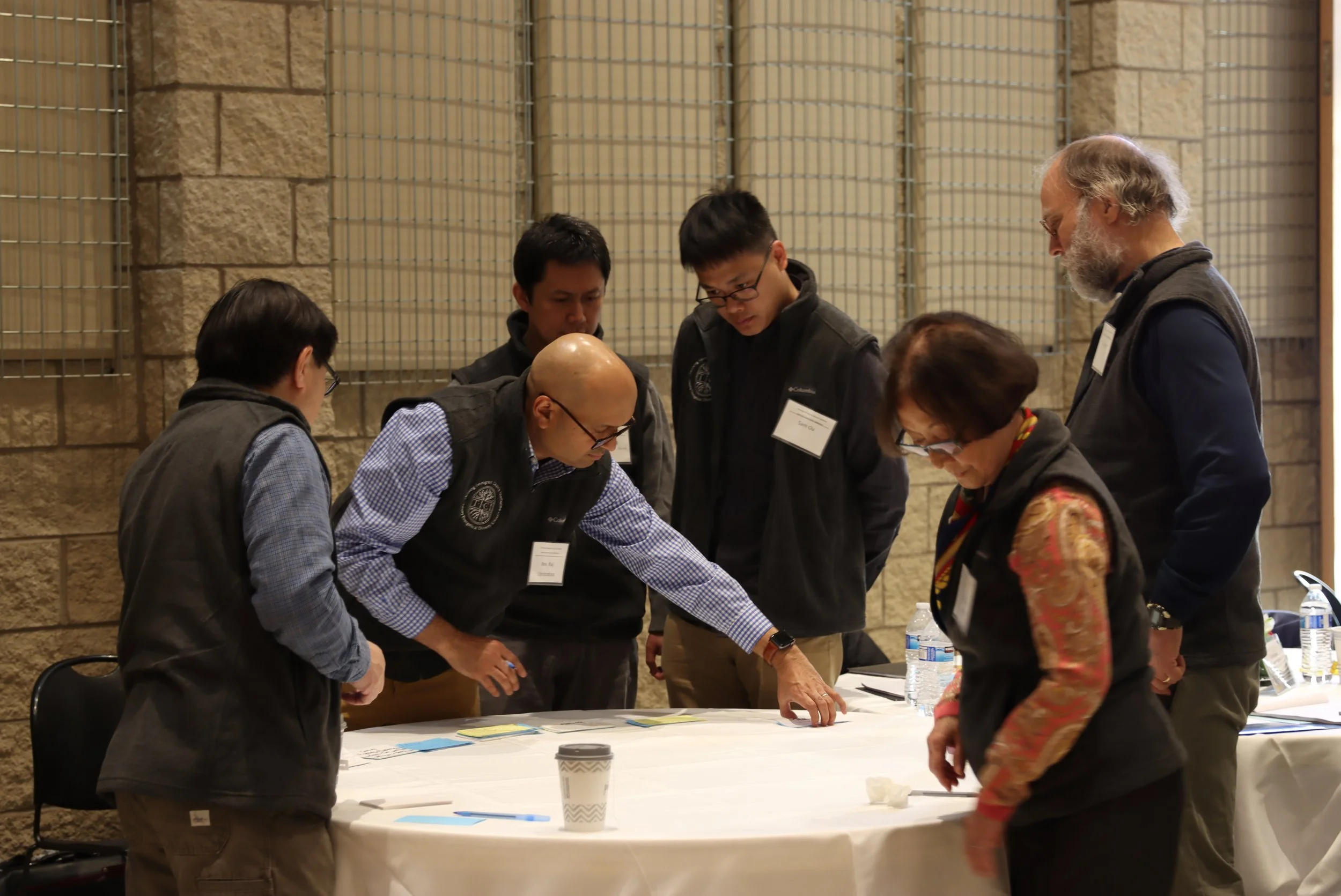 Group of six people gathered around a table, engaged in discussion, in an indoor setting with brick and metal mesh walls.