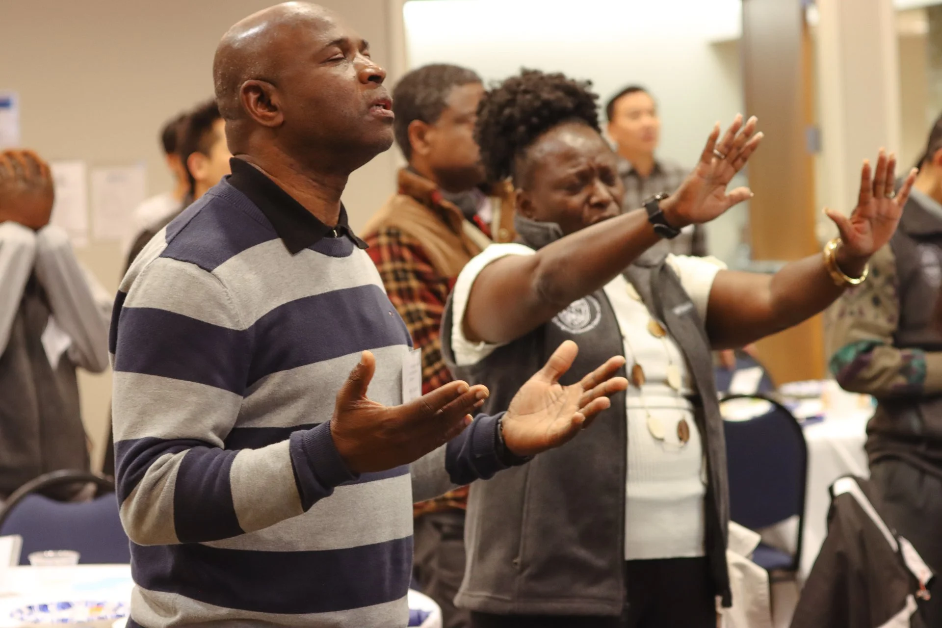 Group of people standing in a prayerful or meditative pose, with eyes closed and hands raised or clasped, in an indoor setting.
