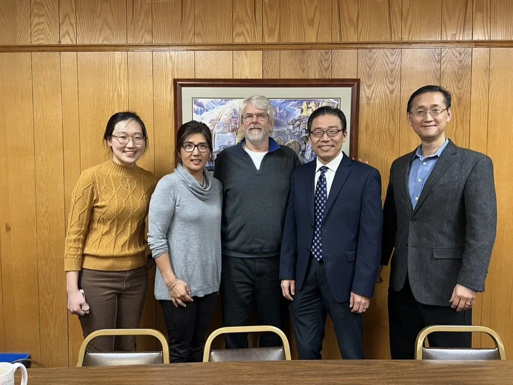 Four Korean individuals and one Caucasian individual in a group photo in front of wood paneled background with framed art