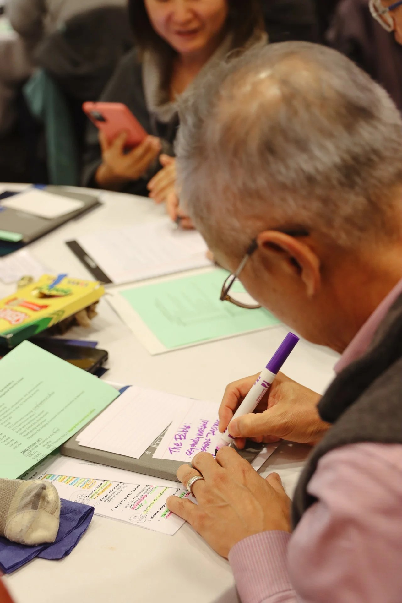 A man with gray hair and glasses writes in a notebook with a purple marker while two women look on, one holding a smartphone, at a table cluttered with papers, brochures, and snacks.