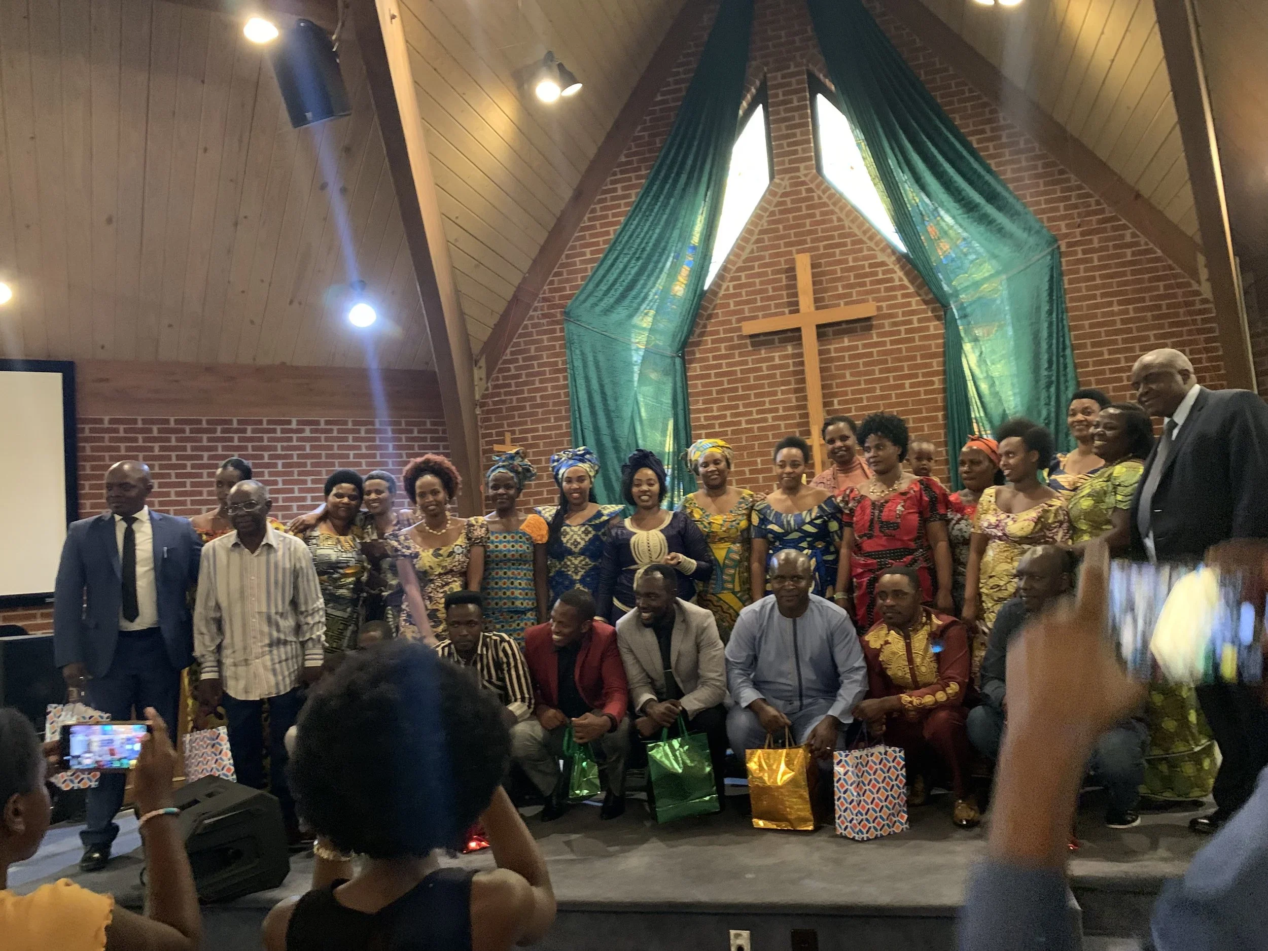 African church members at the front of a church santuary with a brick wall background and a wooden cross in the center