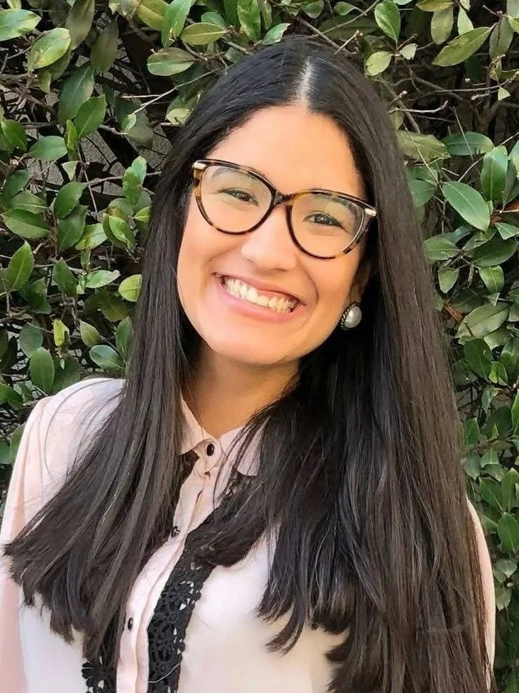 A young woman with long dark hair wearing glasses and pearl earrings, smiling, standing in front of green leafy bushes.