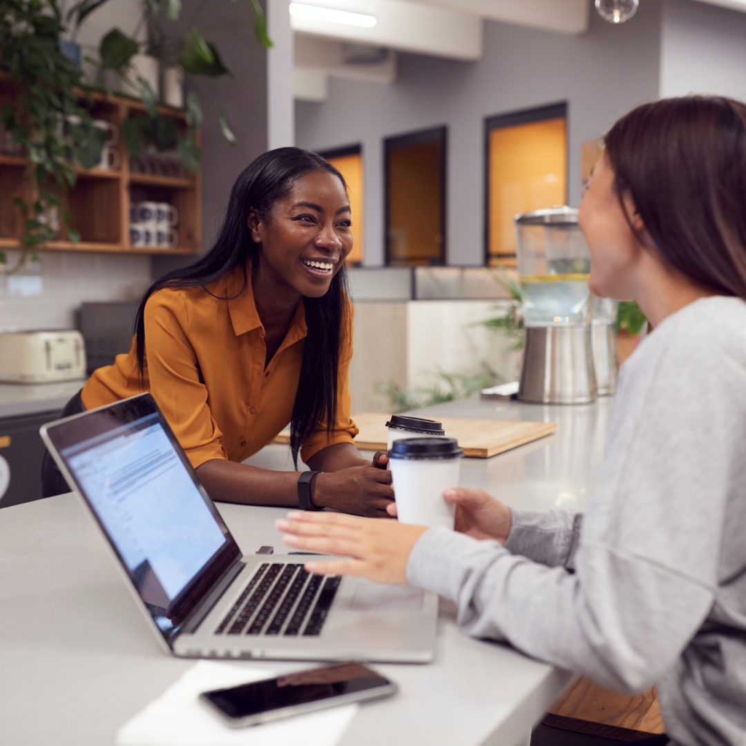 happy woman leaning over smiling and having a conversation with another woman who is using a laptop and drinking coffee