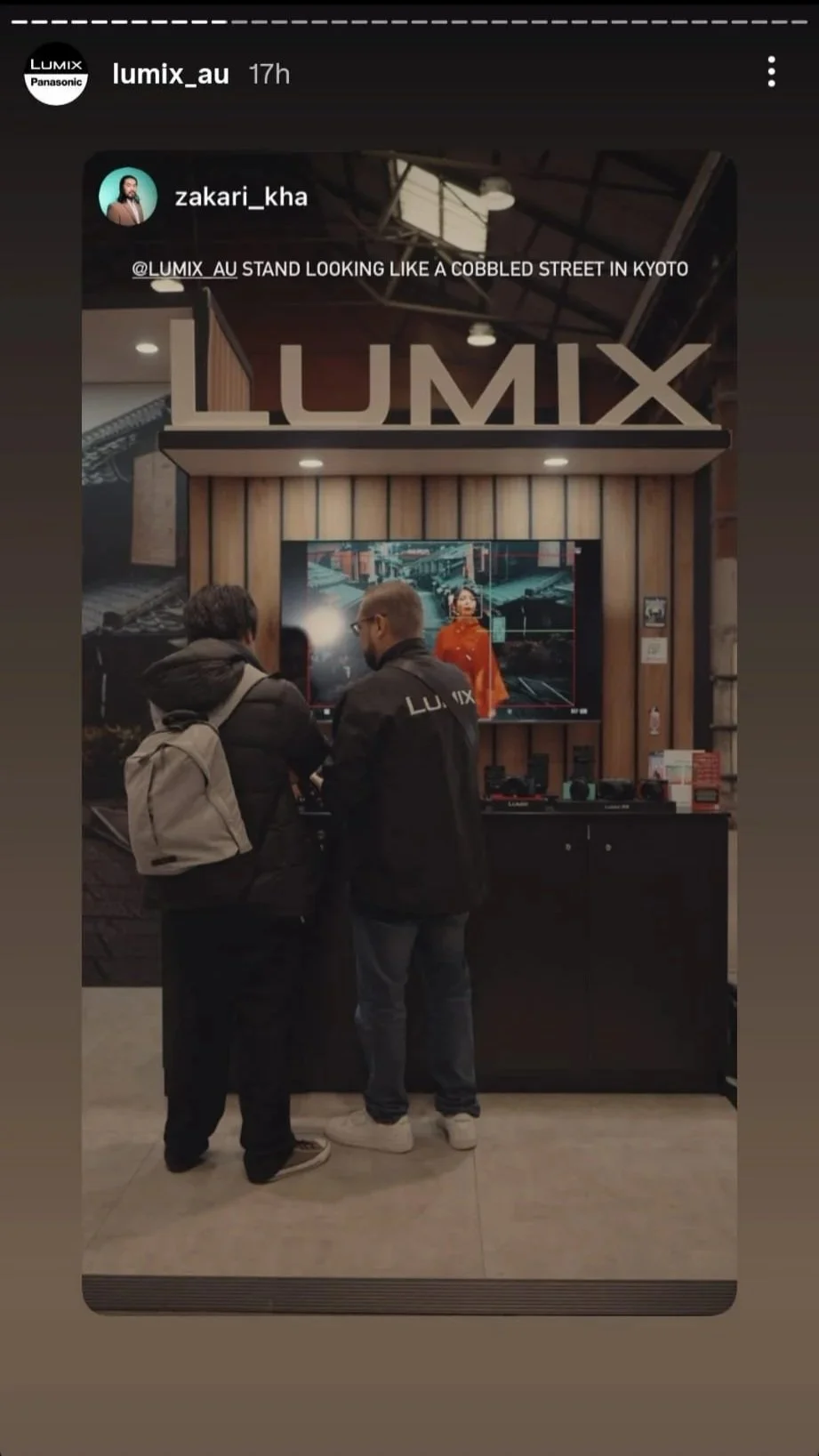 Two people standing in front of a digital display at a LUMIX booth in an indoor venue, with a large LUMIX sign above and a poster of a woman in an orange outfit on the display.