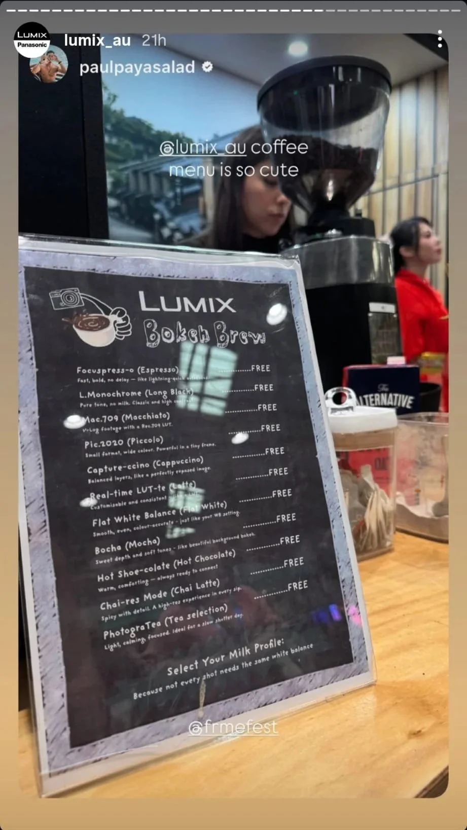 A menu titled 'Lumix Bokah Brew' with various coffee options listed, displayed on a clear stand at a coffee shop, with a coffee grinder in the background and two women, one in black and one in red, visible behind the counter.