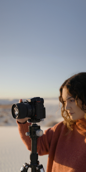Woman adjusting a camera on a tripod outdoors with a sandy landscape and clear sky in the background.