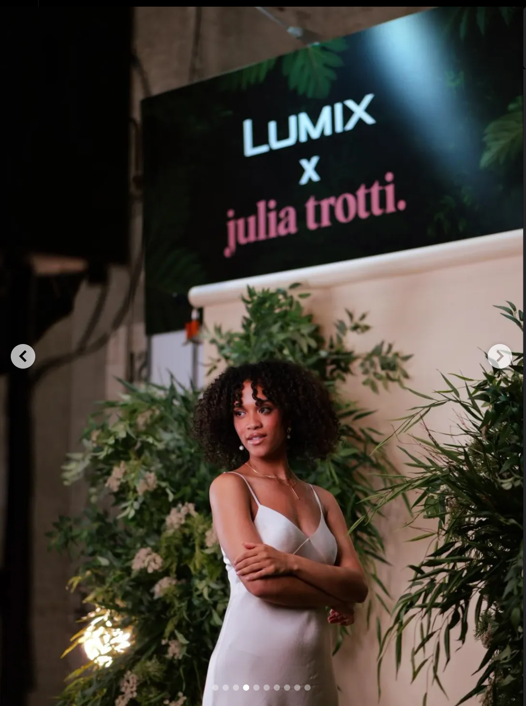 A woman with curly hair wearing a white dress, standing in front of lush greenery, at a LUMIX x Julia Trotti event.