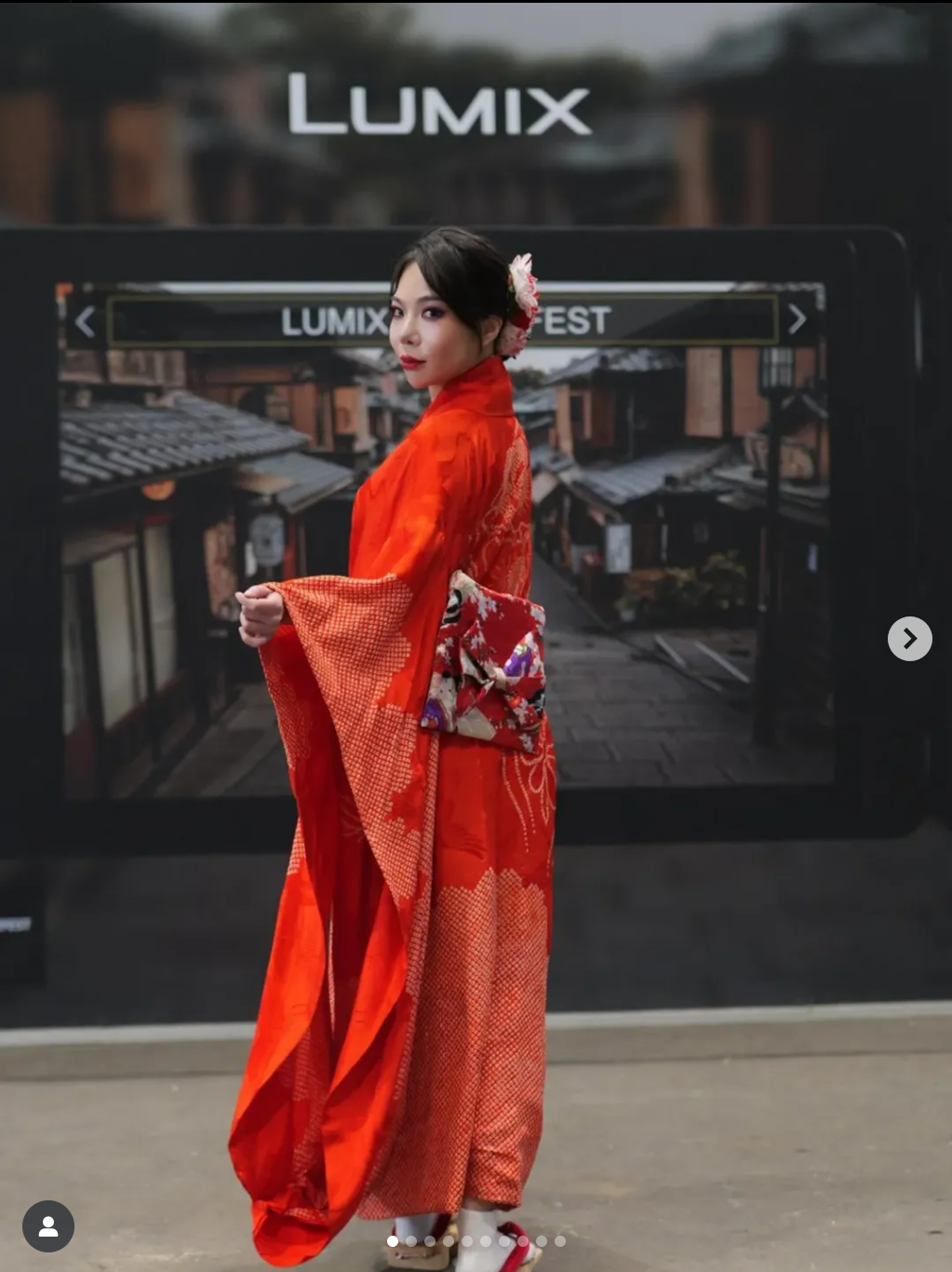 A woman dressed in a traditional red kimono stands in front of a large camera display that reads "LUMIX FEST." She has a floral hair accessory and is posing with a traditional Japanese hairstyle and makeup, with a background resembling a Japanese str