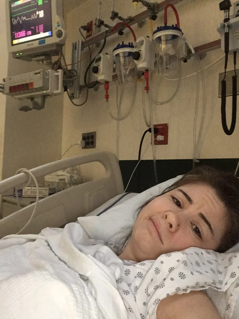 woman in hospital bed with IV bags and monitors, looking toward the camera