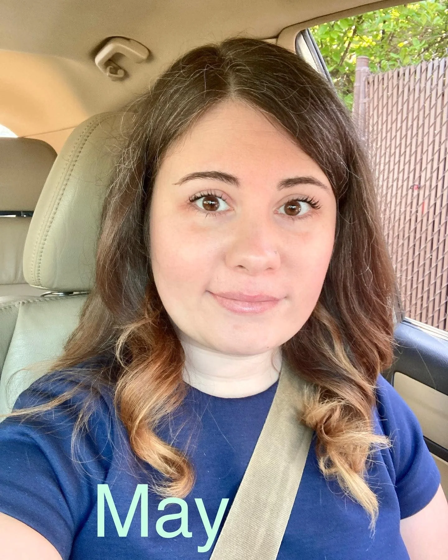 woman in a navy shirt taking a car selfie, labeled “May,” with trees and a fence outside the window