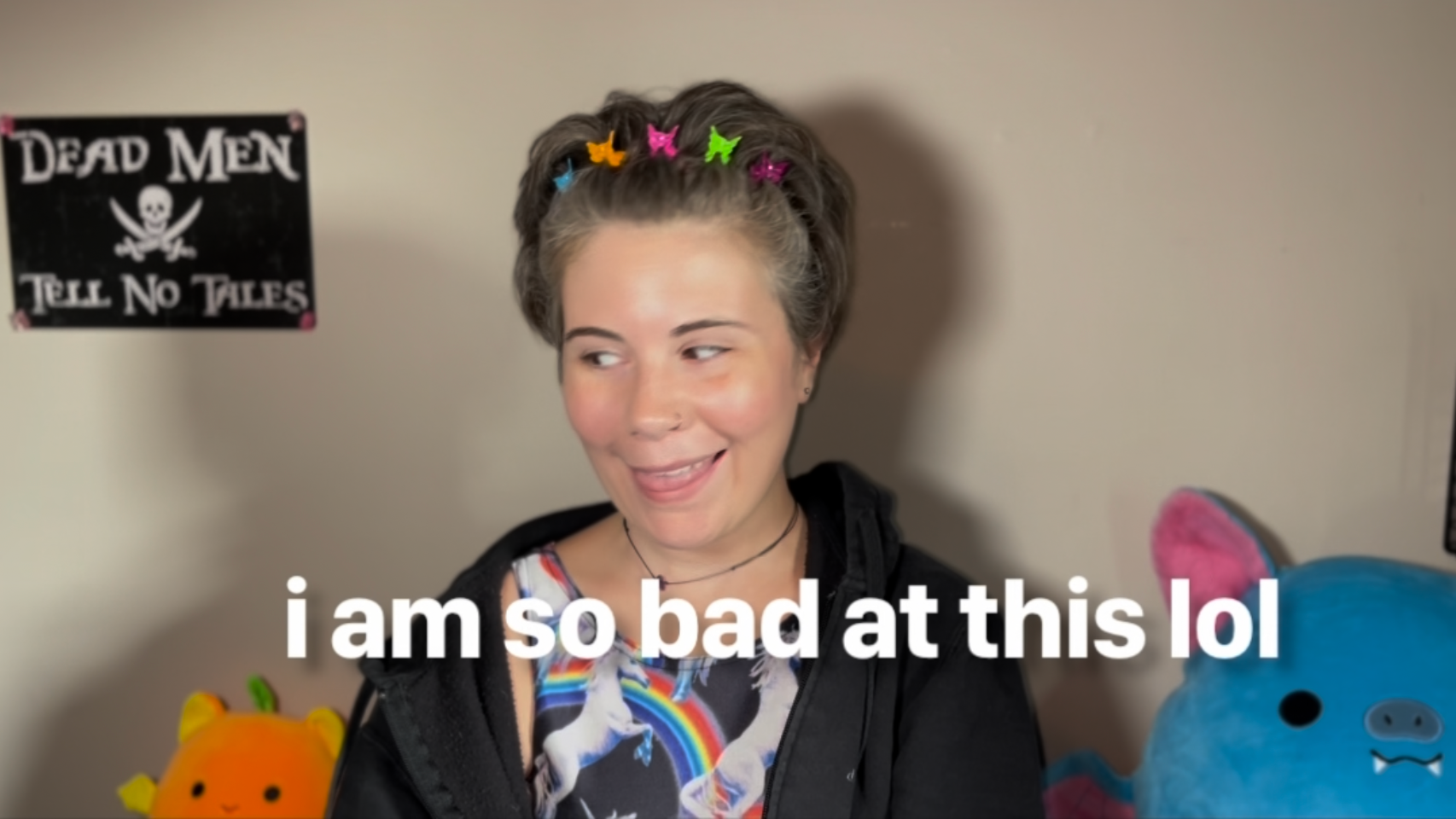 woman smiling in front of beige wall, wearing colorful butterfly clips and a unicorn-print shirt with text overlay: “i am so bad at this lol”