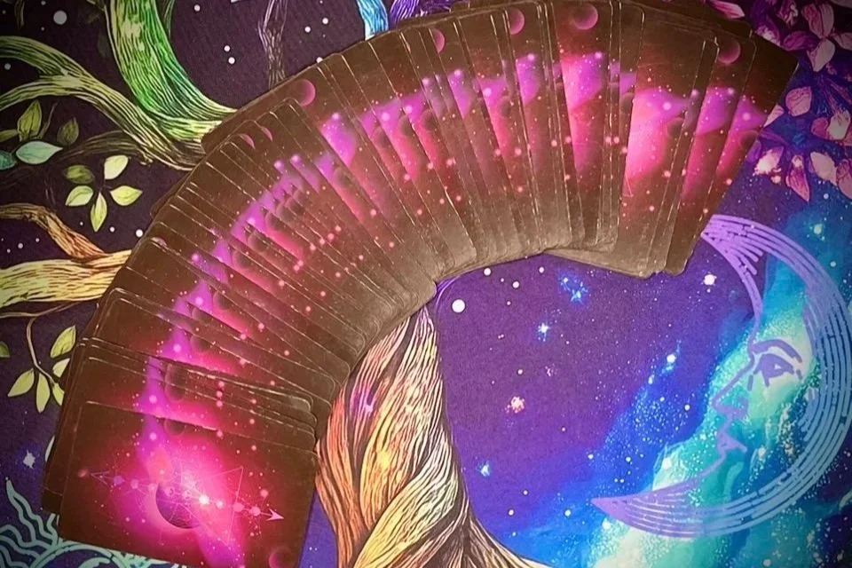 a full tarot deck fanned out in a wide arc over a vivid, galactic background with swirling stars and cosmic imagery in pink, gold, and blue tones.