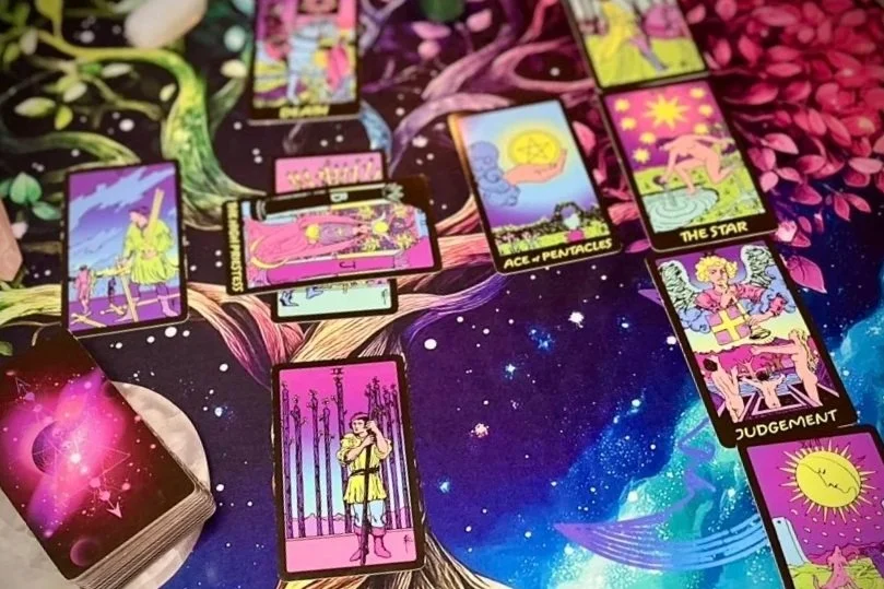 several tarot cards laid out face up on a vibrant, cosmic-patterned cloth, featuring bold colors and galactic imagery. visible cards include the star, ace of pentacles, and judgment. the deck sits off to the left.