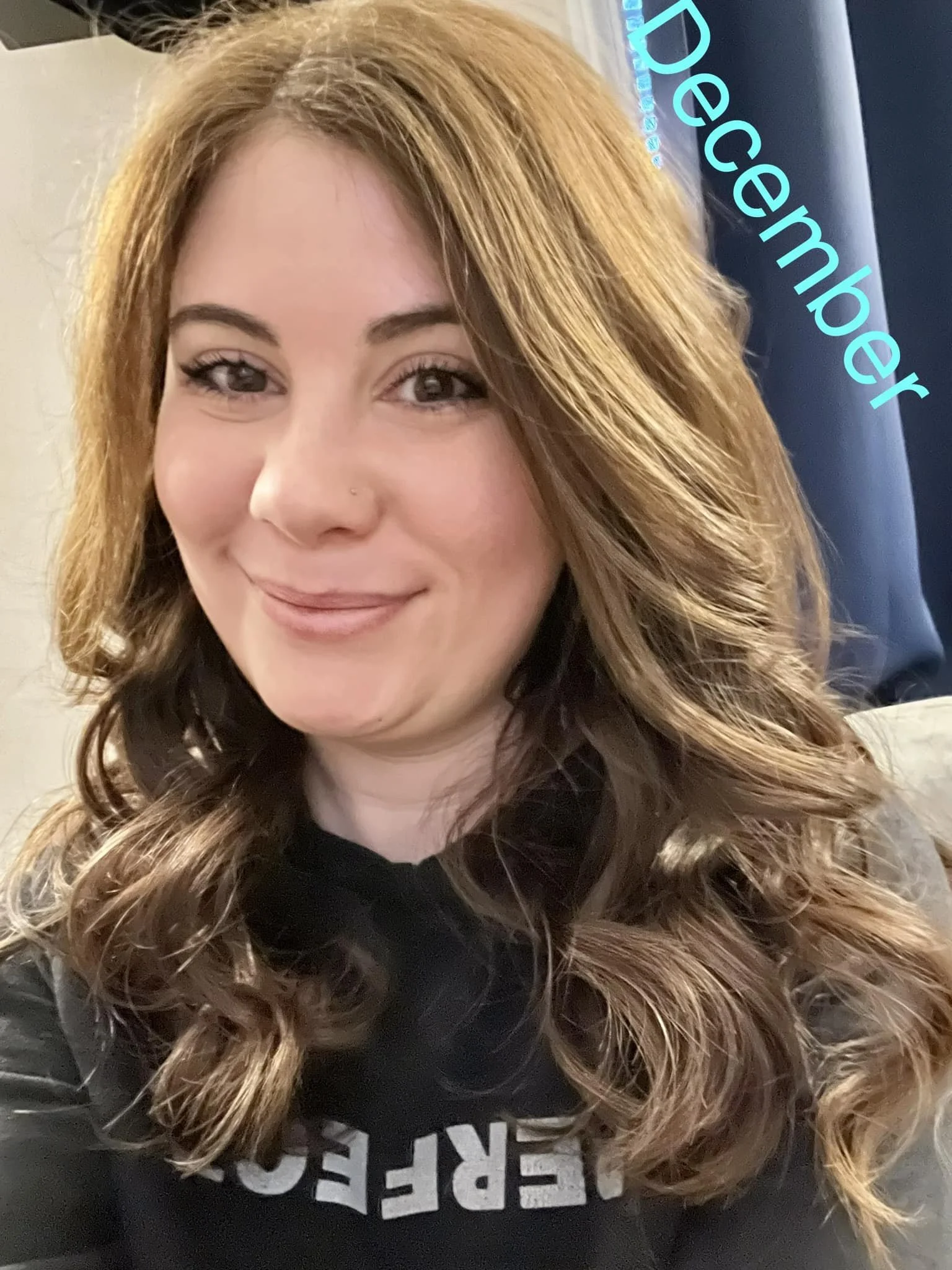 woman with long curled brunette hair smiling at the camera in soft indoor lighting, wearing a black sweatshirt with partial white lettering, with the word ‘December’ written in teal text overlay