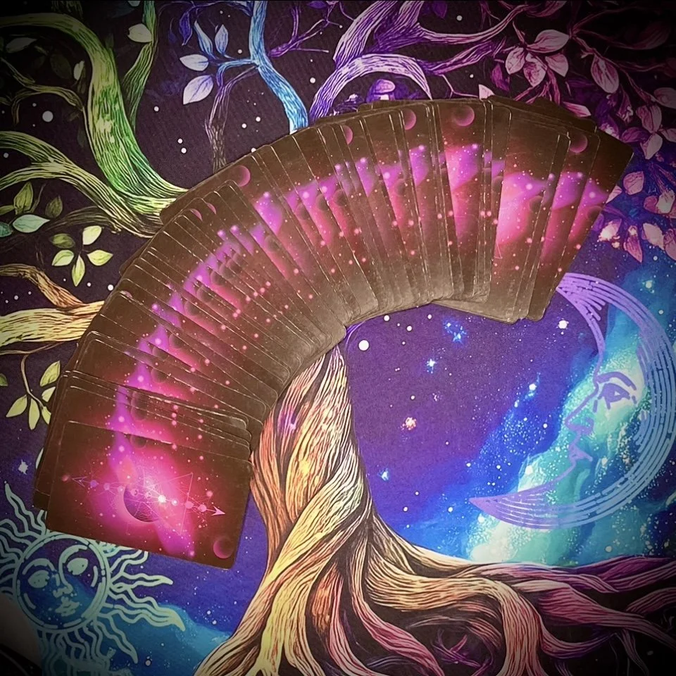a full tarot deck fanned out in a wide arc over a vivid, galactic background with swirling stars and cosmic imagery in pink, gold, and blue tones.