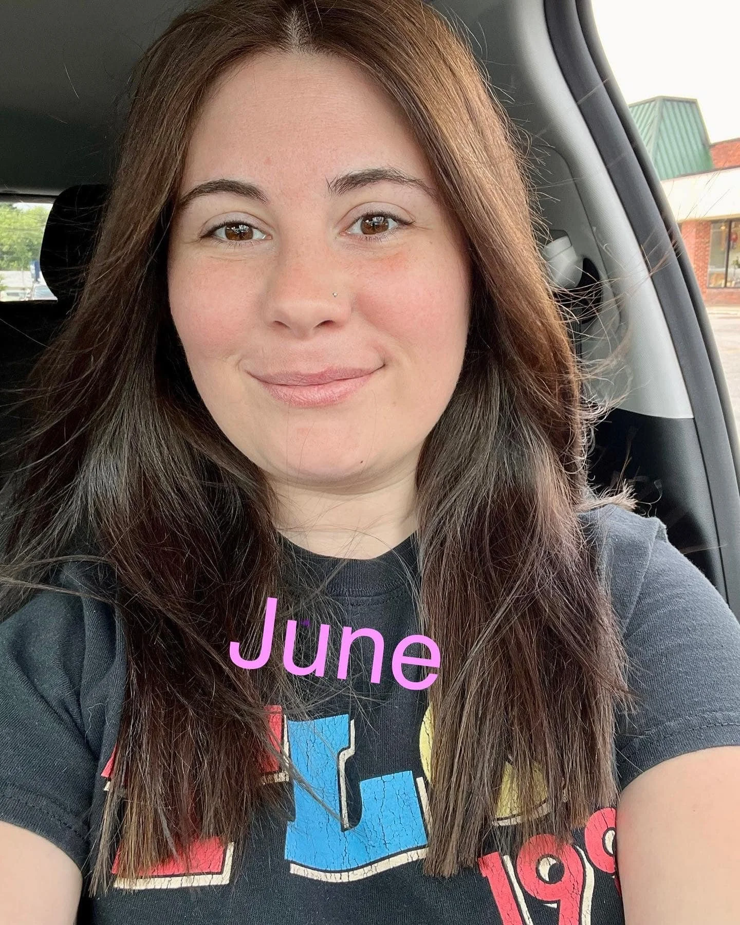 brunette woman in a car smiling with "June" written in pink overlay text, buildings and trees visible outside the window