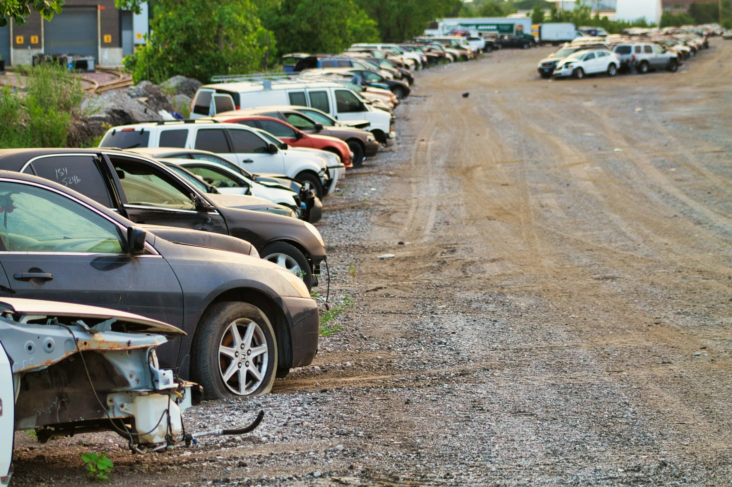 A parking lot with many damaged and wrecked cars lined up on a dirt surface, some missing parts, and others appearing intact.