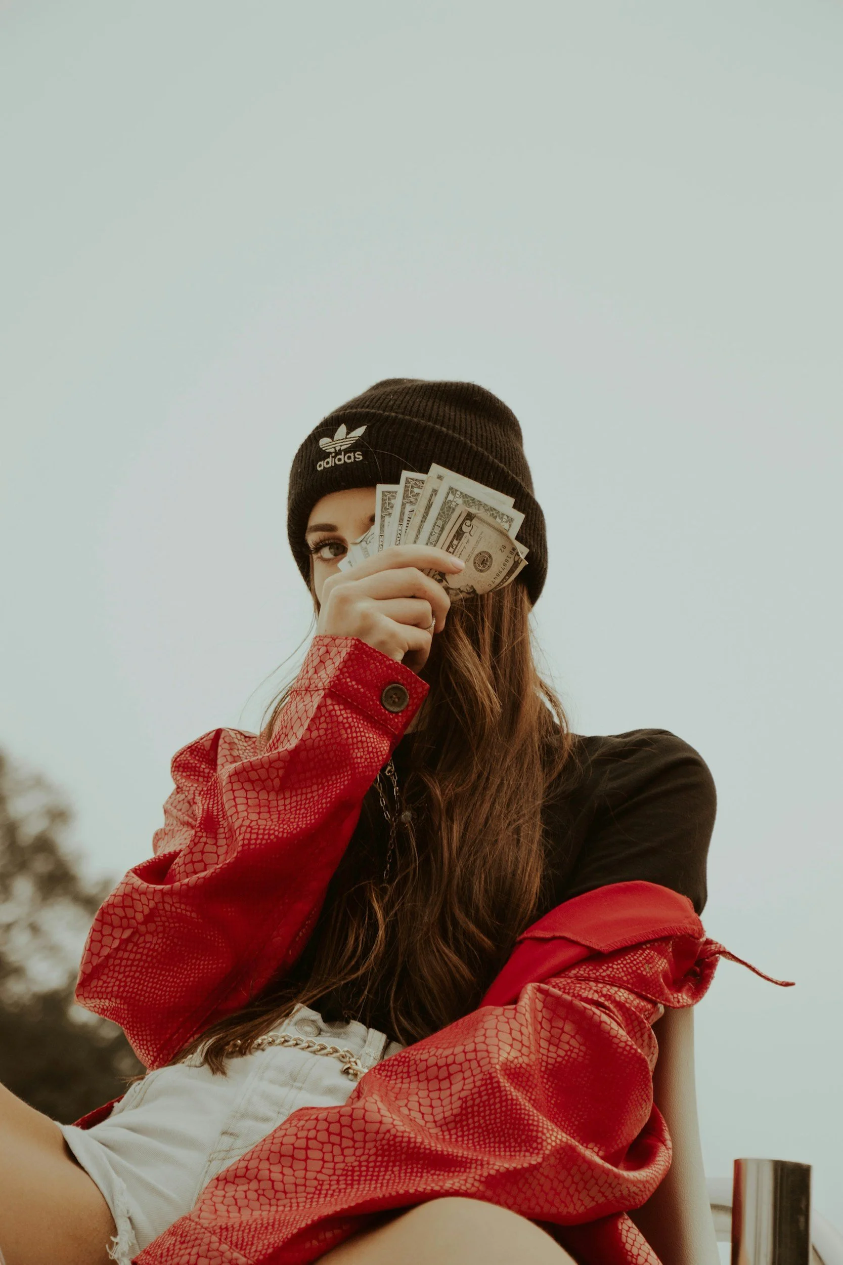 A woman dressed in a red jacket with snake pattern, wearing a black Adidas beanie, holds a fan of U.S. dollar bills over her face, sitting outdoors.