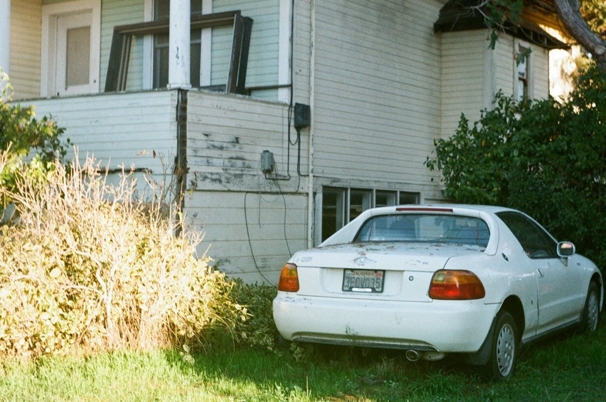 A white, weathered two-door car parked on a grassy area beside an old, dilapidated house with beige siding, small windows, and a broken porch. Bushes and trees are nearby.
