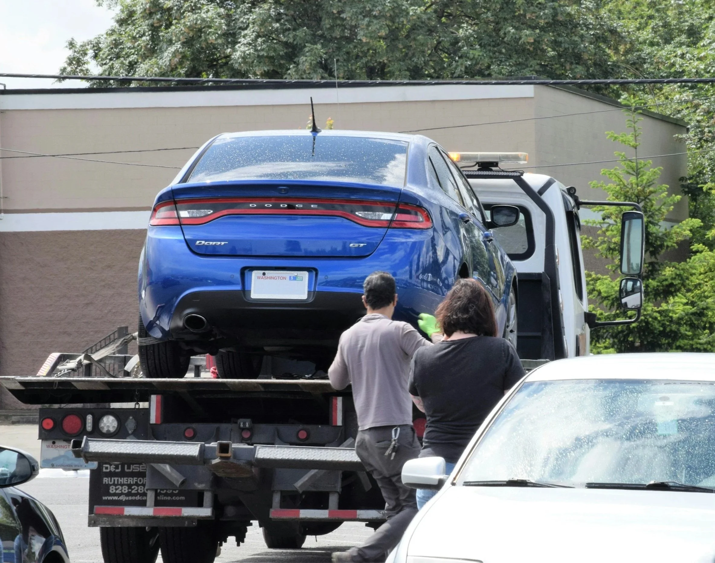 A blue Dodge Dart GT car being loaded onto a flatbed tow truck, with two people inspecting or working on it, in a parking lot near trees and a building.