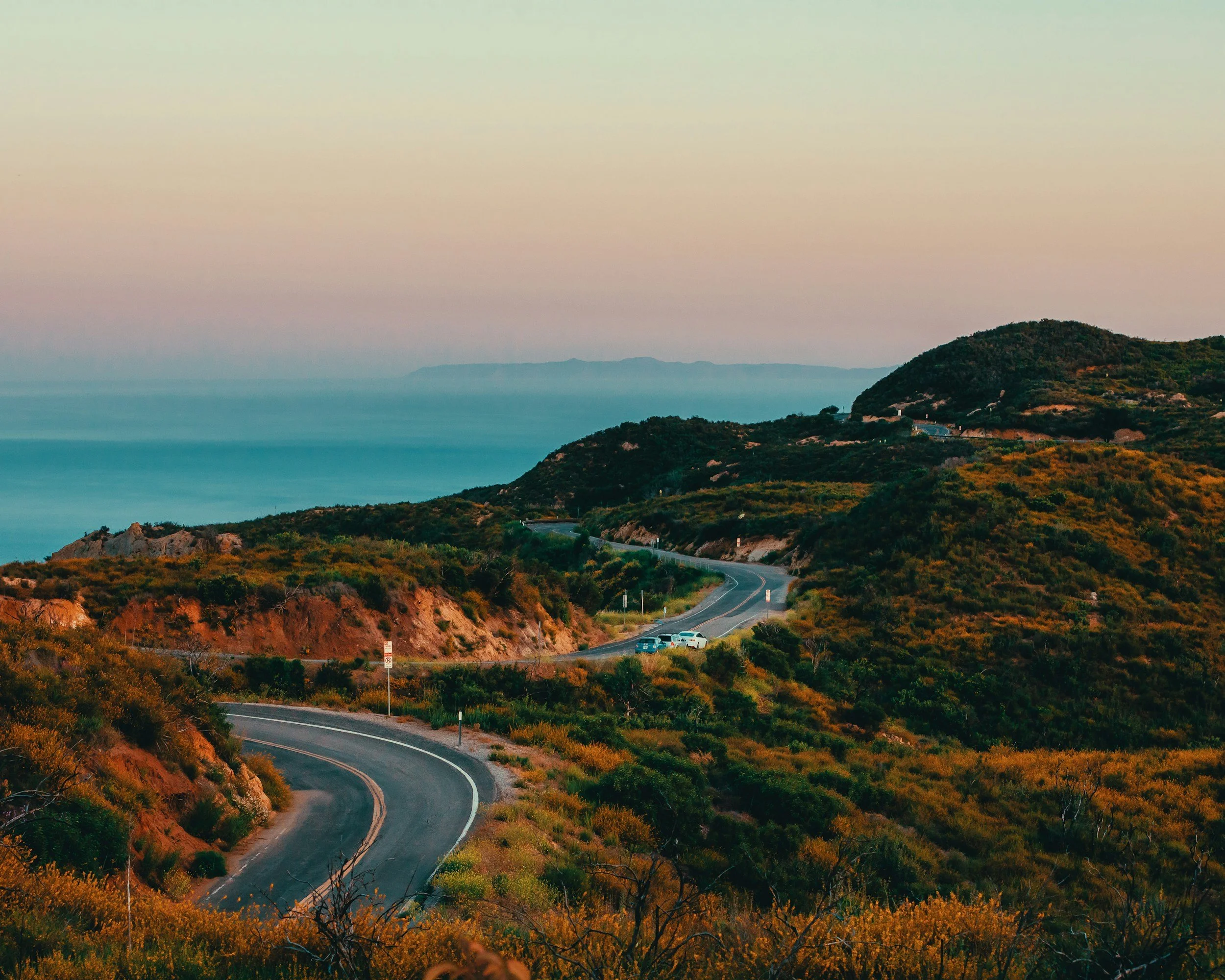Winding coastal road through lush green hills with a view of the ocean and distant mountains during sunset.