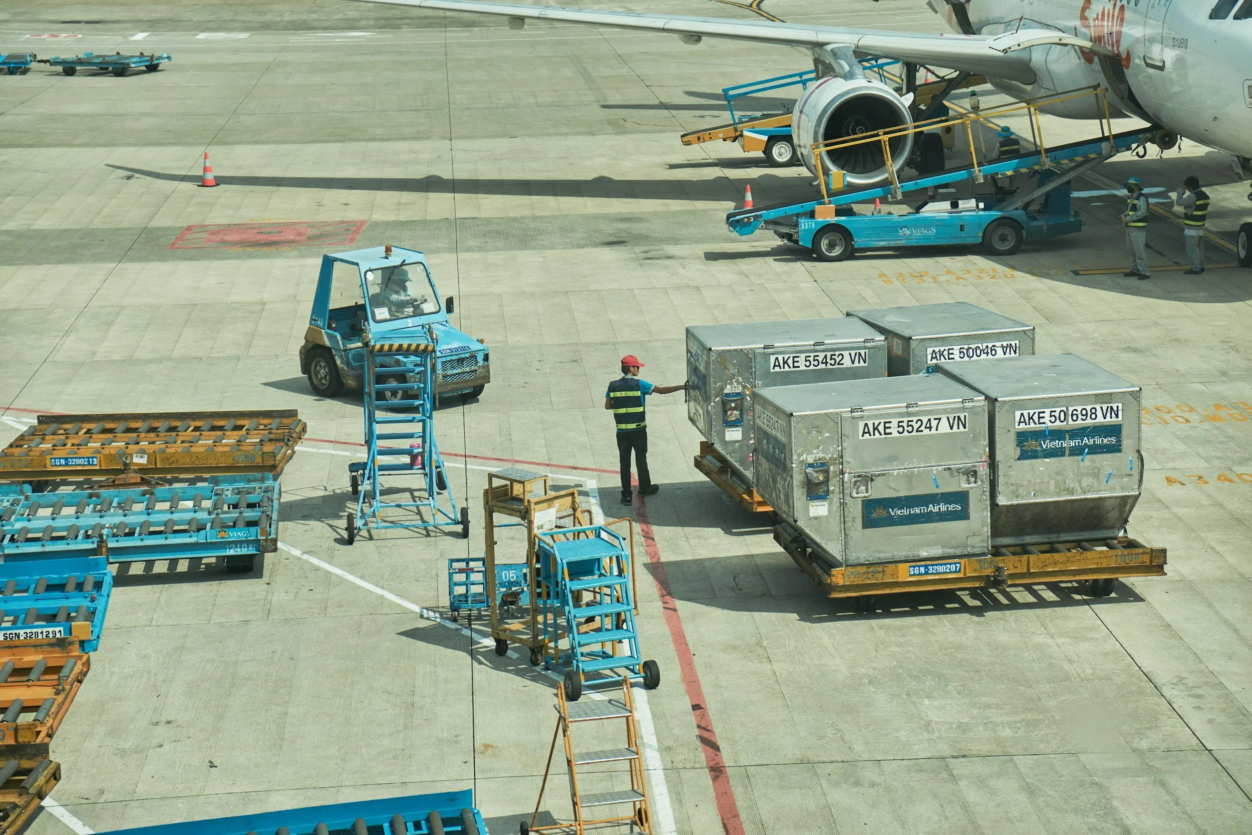 At an airport, luggage carts and cargo containers are on the tarmac near an airplane. Ground crew members are handling luggage and cargo, including various labeled containers. Some staff are near the plane's cargo hold, and others are maneuvering carts and equipment.