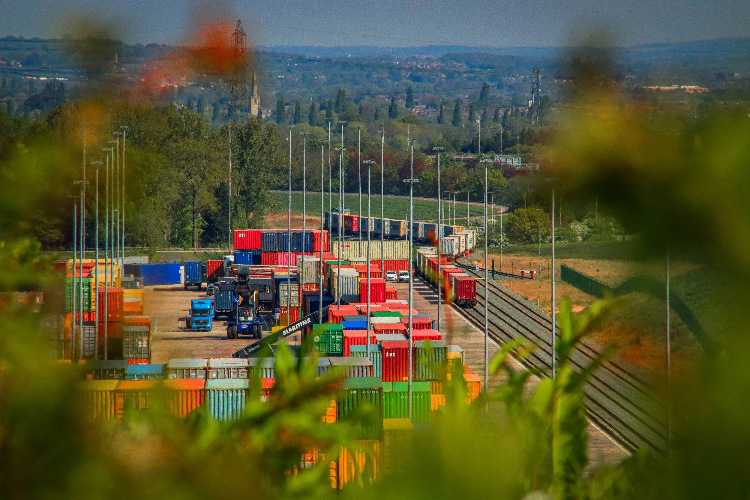 View of a freight container yard with stacked colorful shipping containers, train tracks with a train, and vehicles in the yard, with green trees and hills in the background.