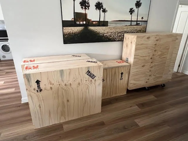 Three large, unfinished wooden boxes or panels leaning against a wall inside a room, with a landscape painting of palm trees and a beach scene hanging above them.