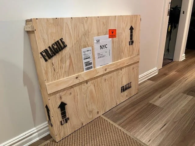 Wooden shipping crate with 'FRAGILE' and 'UP' markings leaning against a white wall inside a house.