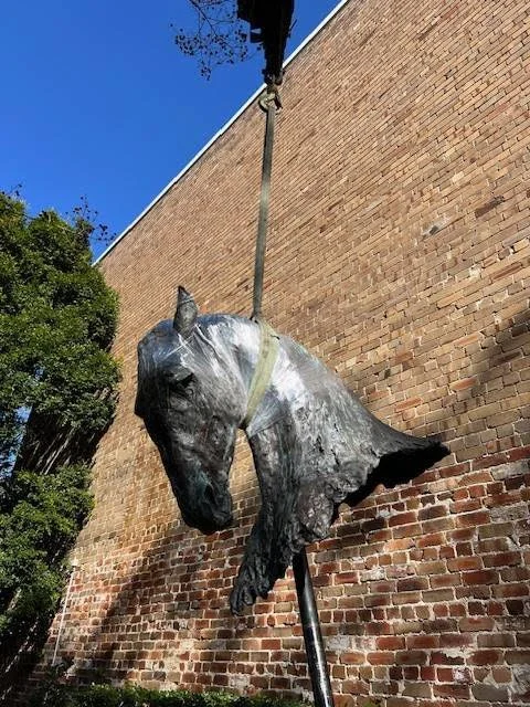 A sculpture of a horse's head and neck mounted on a pole next to a brick wall, with a clear blue sky in the background.