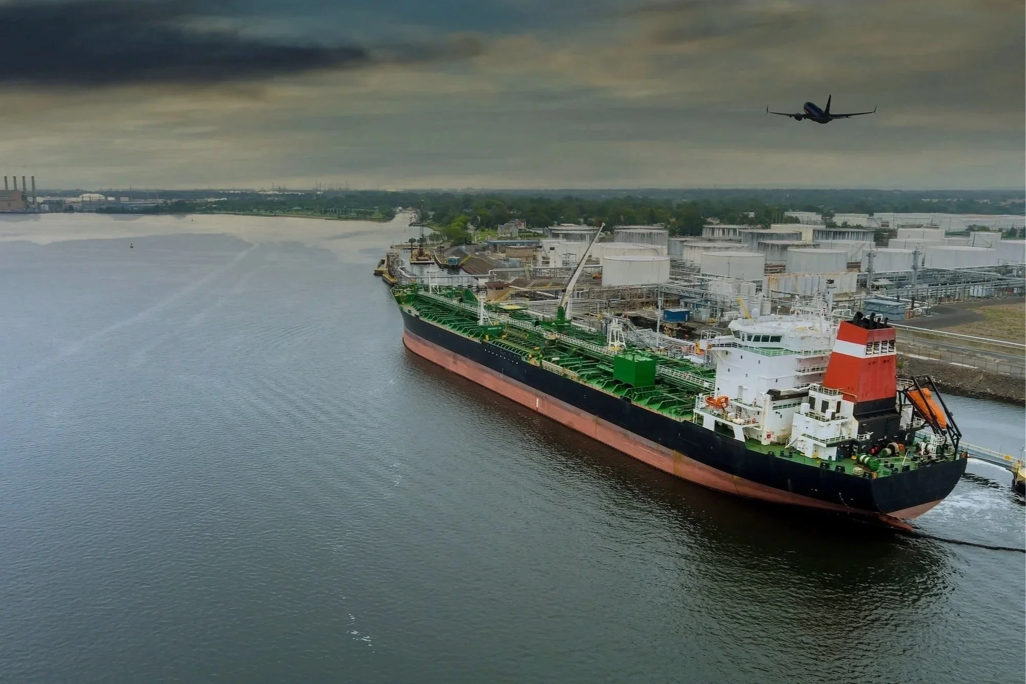 An oil tanker ship passing through a channel with an industrial plant along the shoreline, and a commercial airplane flying overhead in a cloudy sky.