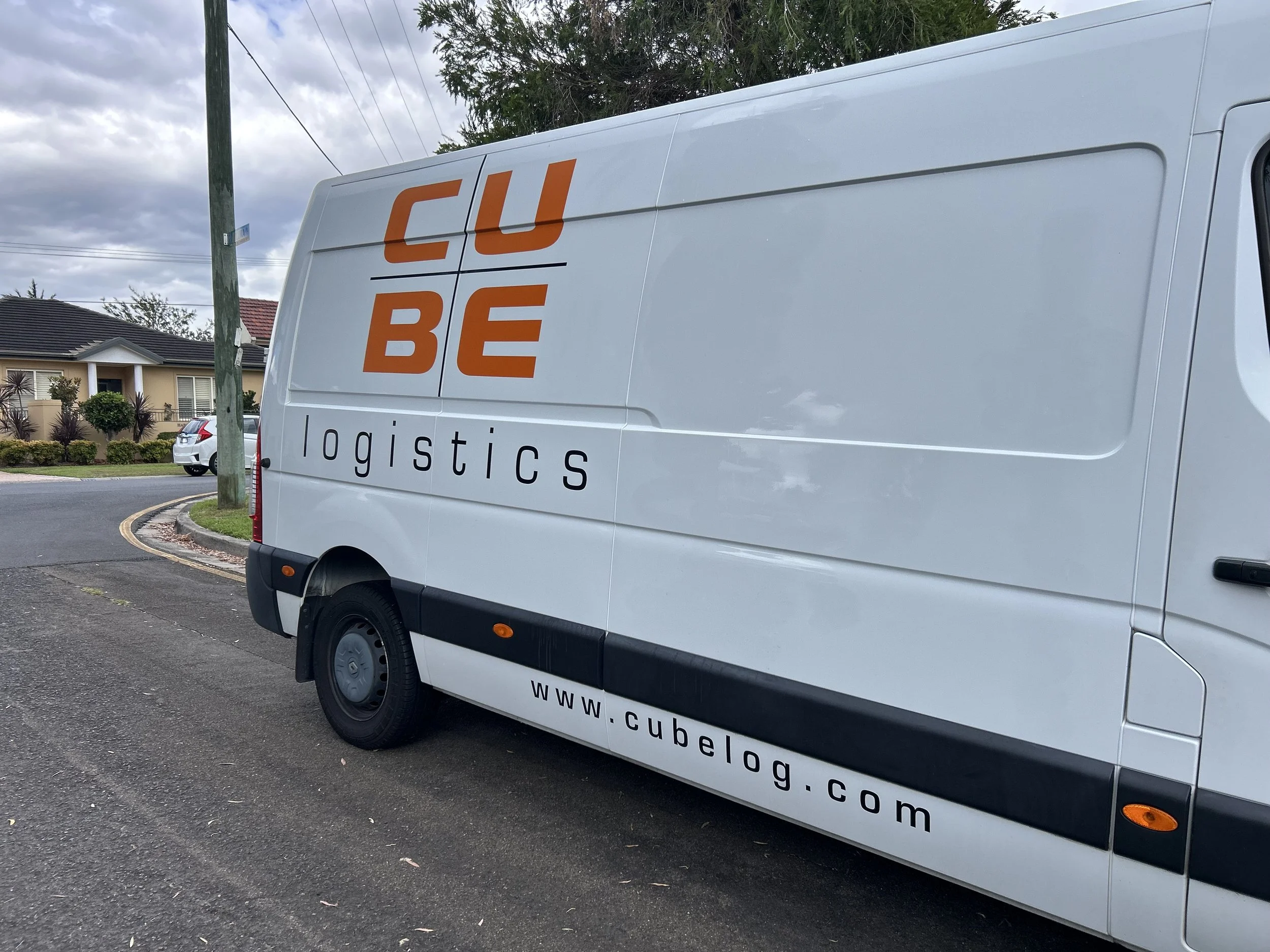 A white delivery van parked on the street with the logo 'CUBE logistics' in orange and black. The website address www.cubelogg.com is visible on the side of the van. There are houses and trees in the background.