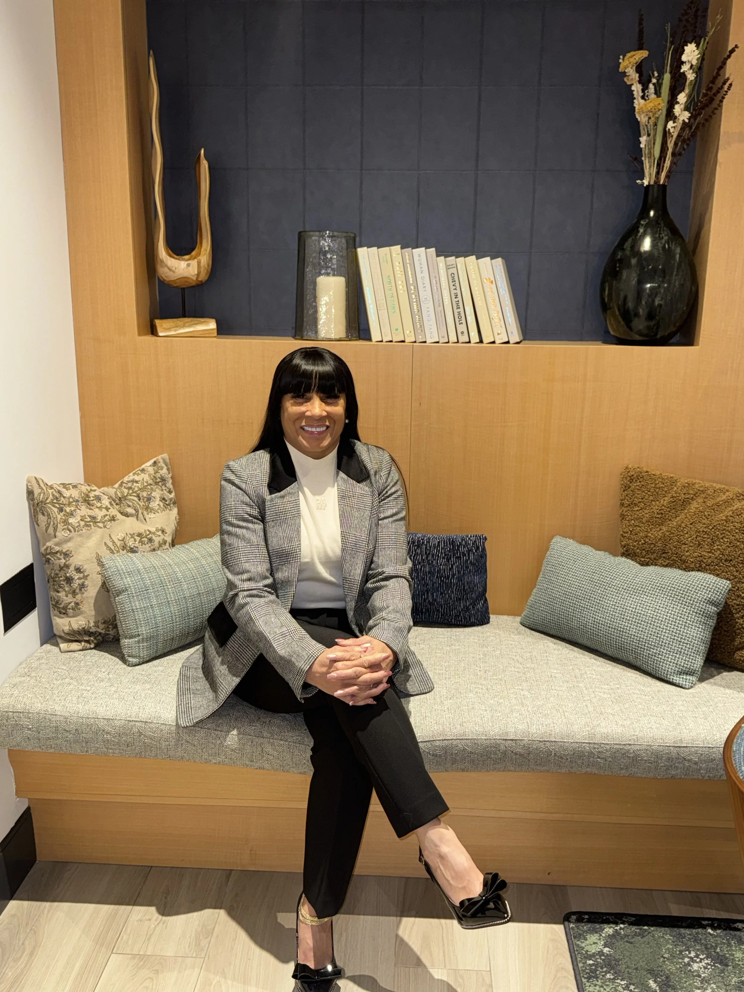 A woman with black hair, wearing a plaid blazer and black pants, sitting on a beige cushioned bench with multiple pillows in a room with wood and dark blue wall decor, smiling at the camera.
