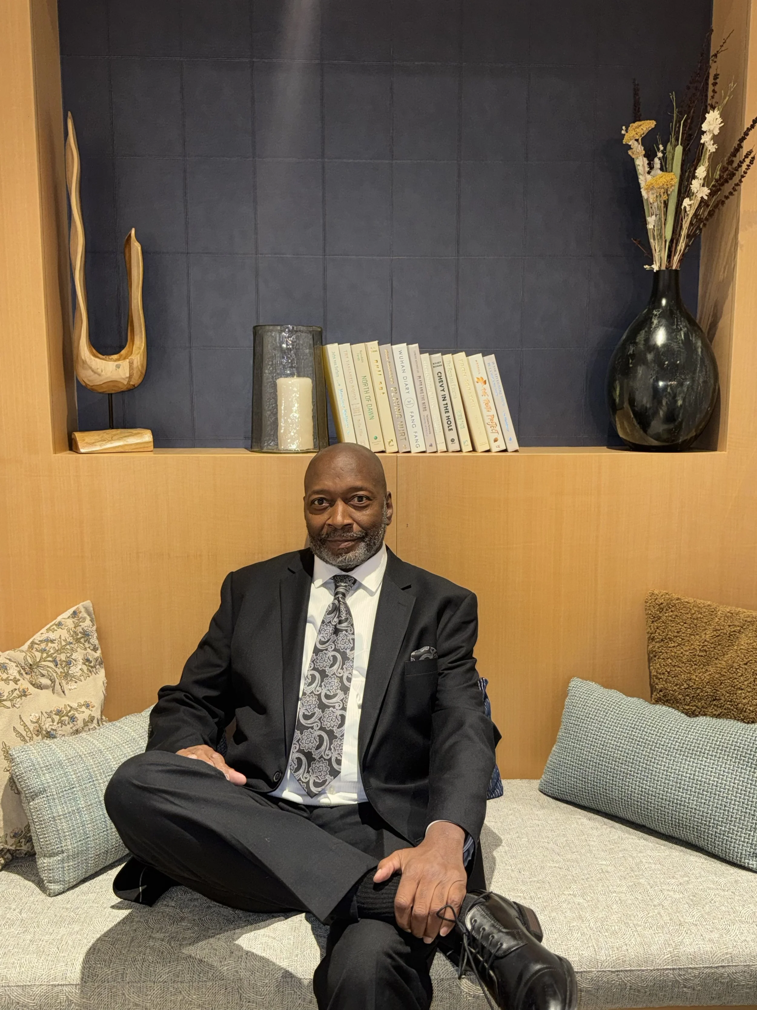 A man dressed in a black suit and tie sitting on a beige cushioned bench with decorative pillows, smiling at the camera.