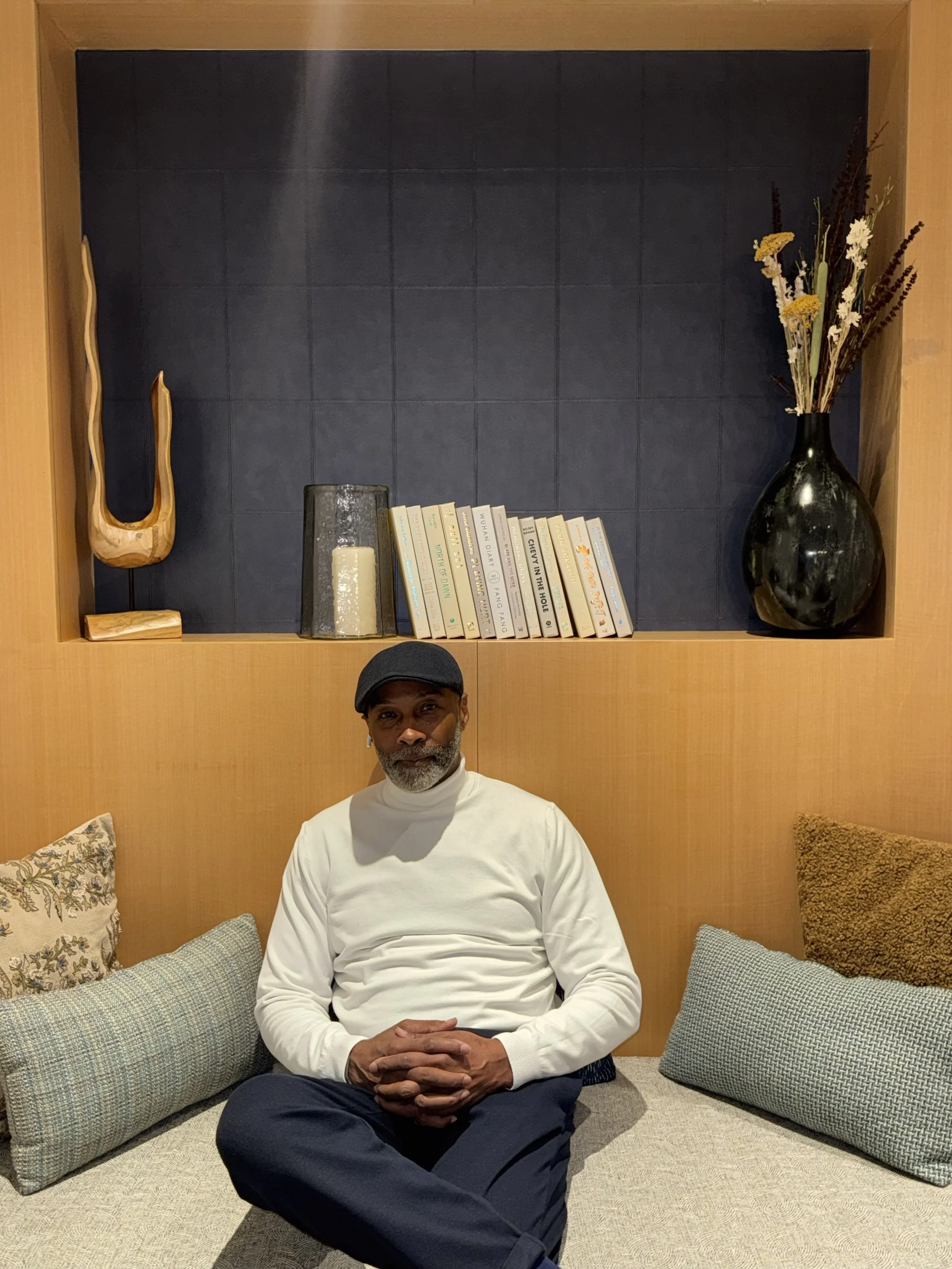 A man wearing a white turtleneck and dark pants, sitting cross-legged on a cushioned bench with decorative pillows, in a room with wood-paneled walls and a built-in shelf featuring books, a black vase with dried flowers, a candle, and an abstract wooden sculpture.
