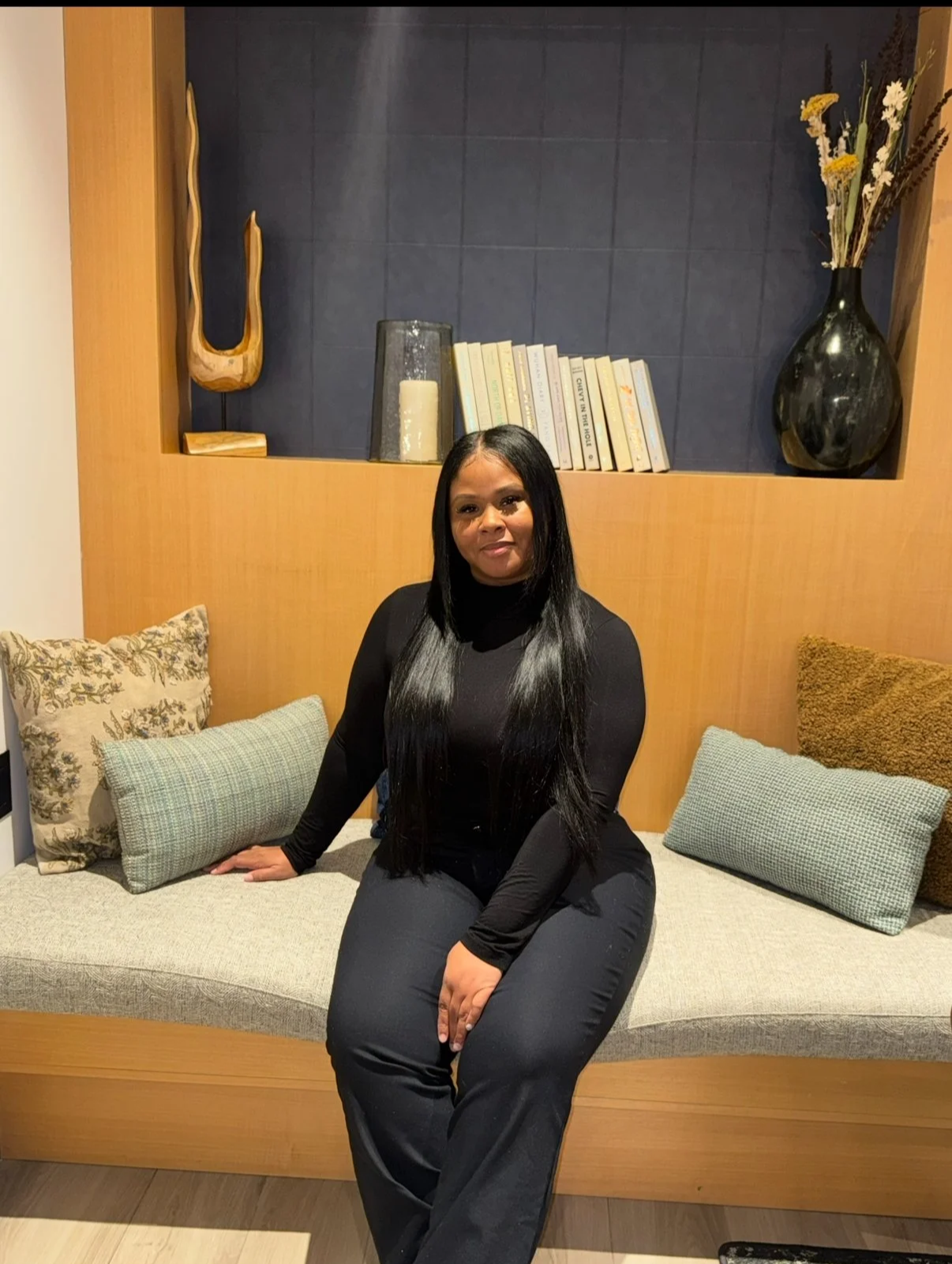 A woman with long black hair sitting on a bench with cushions, wearing a black long sleeve top and black pants, in a room with wooden and blue wall decorations, including books and large vases with dried flowers.