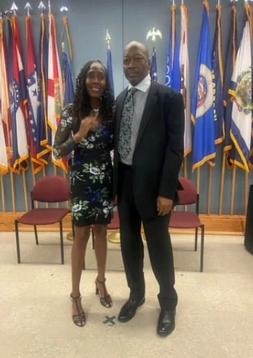 A woman and a man standing together in front of a wall of flags.
The woman is smiling, dressed in a black floral dress and high heels.
The man is wearing a dark suit with a patterned tie, and has a serious expression.