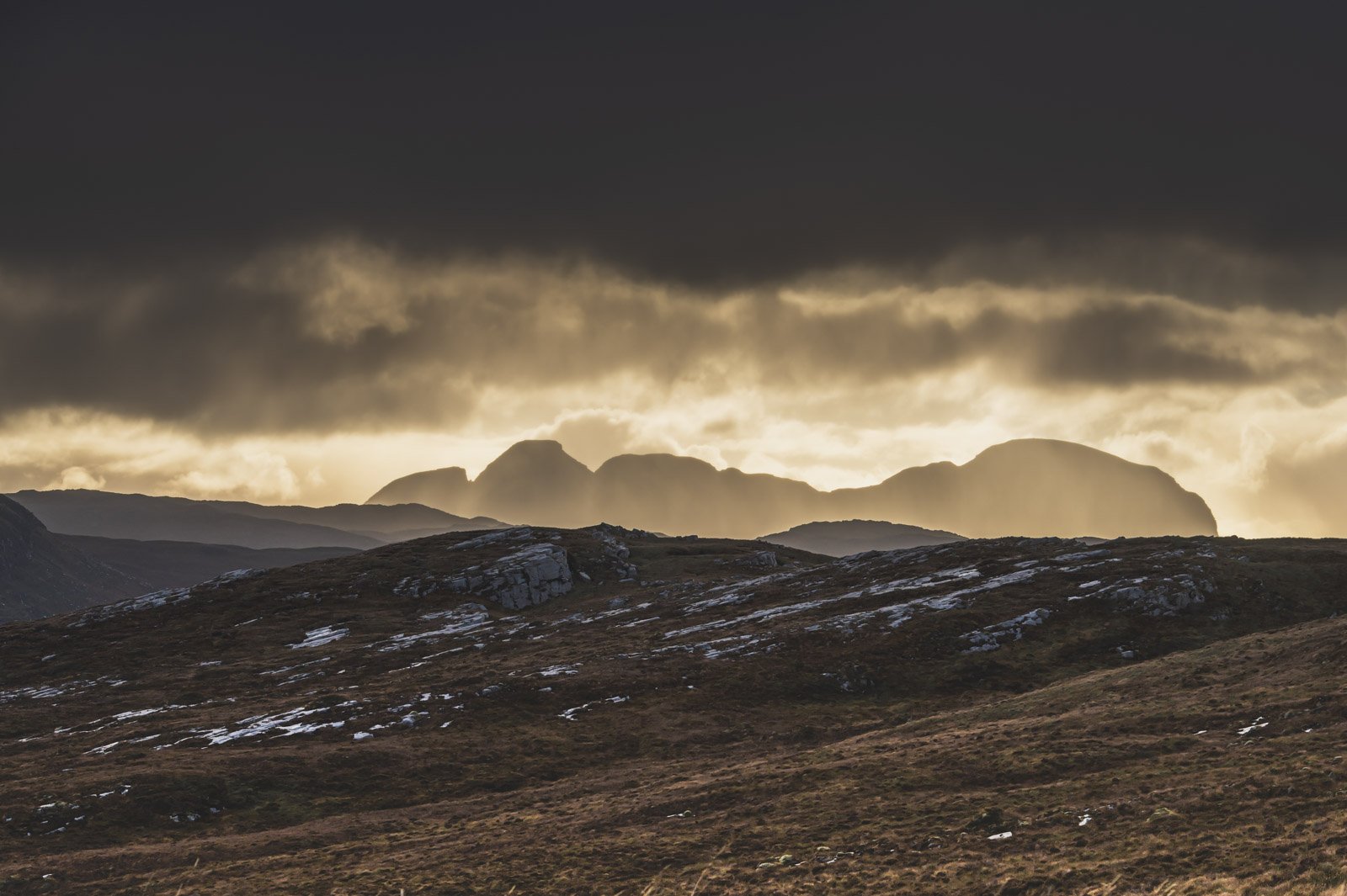 Mountain landscape at sunset with dark clouds overhead and patches of snow on the ground.