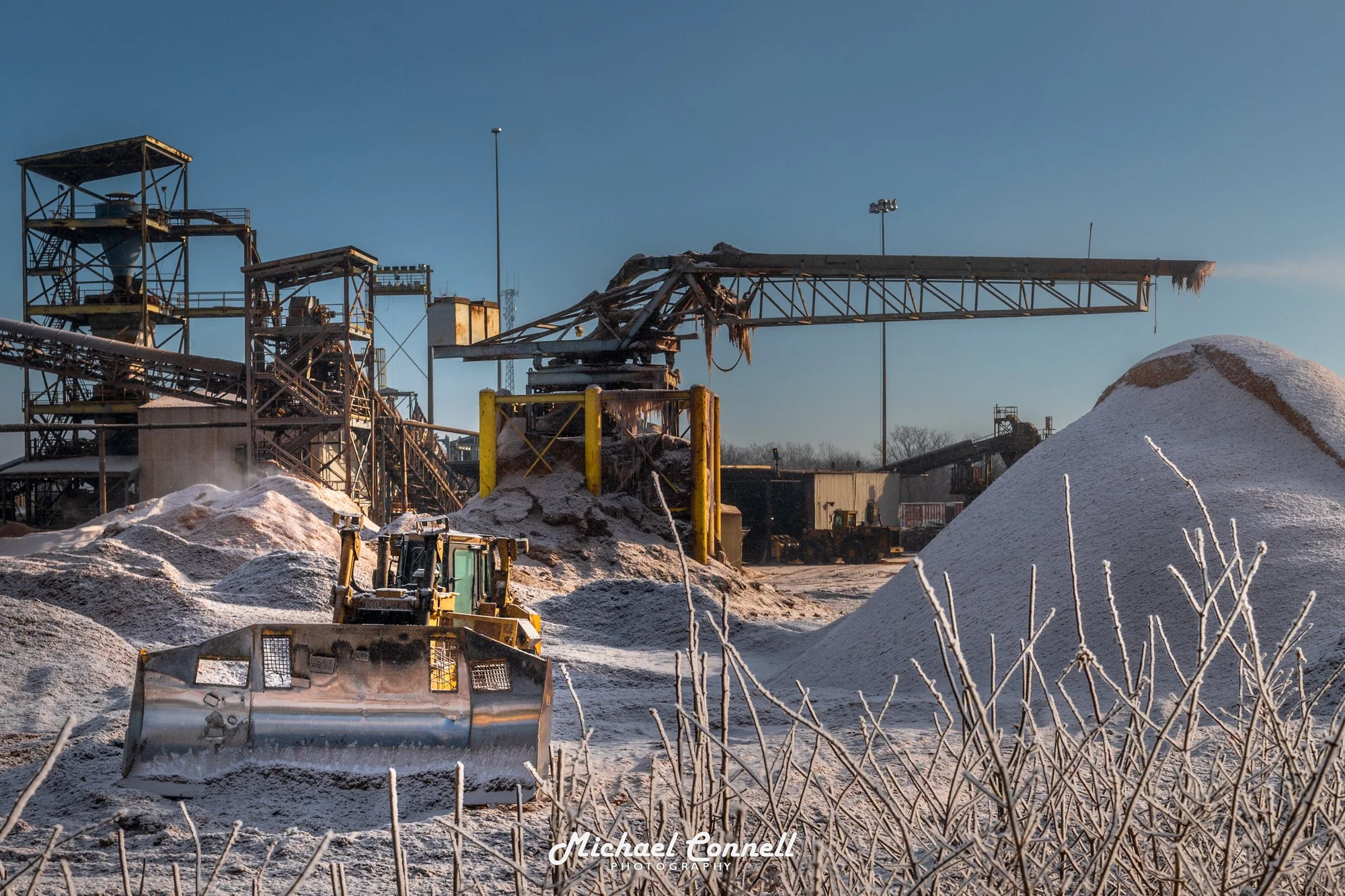 A snow-covered industrial mining site with a yellow bulldozer in the foreground, piles of salt or minerals, and rusted metal structures including a crane and conveyor belts under a clear blue sky.
