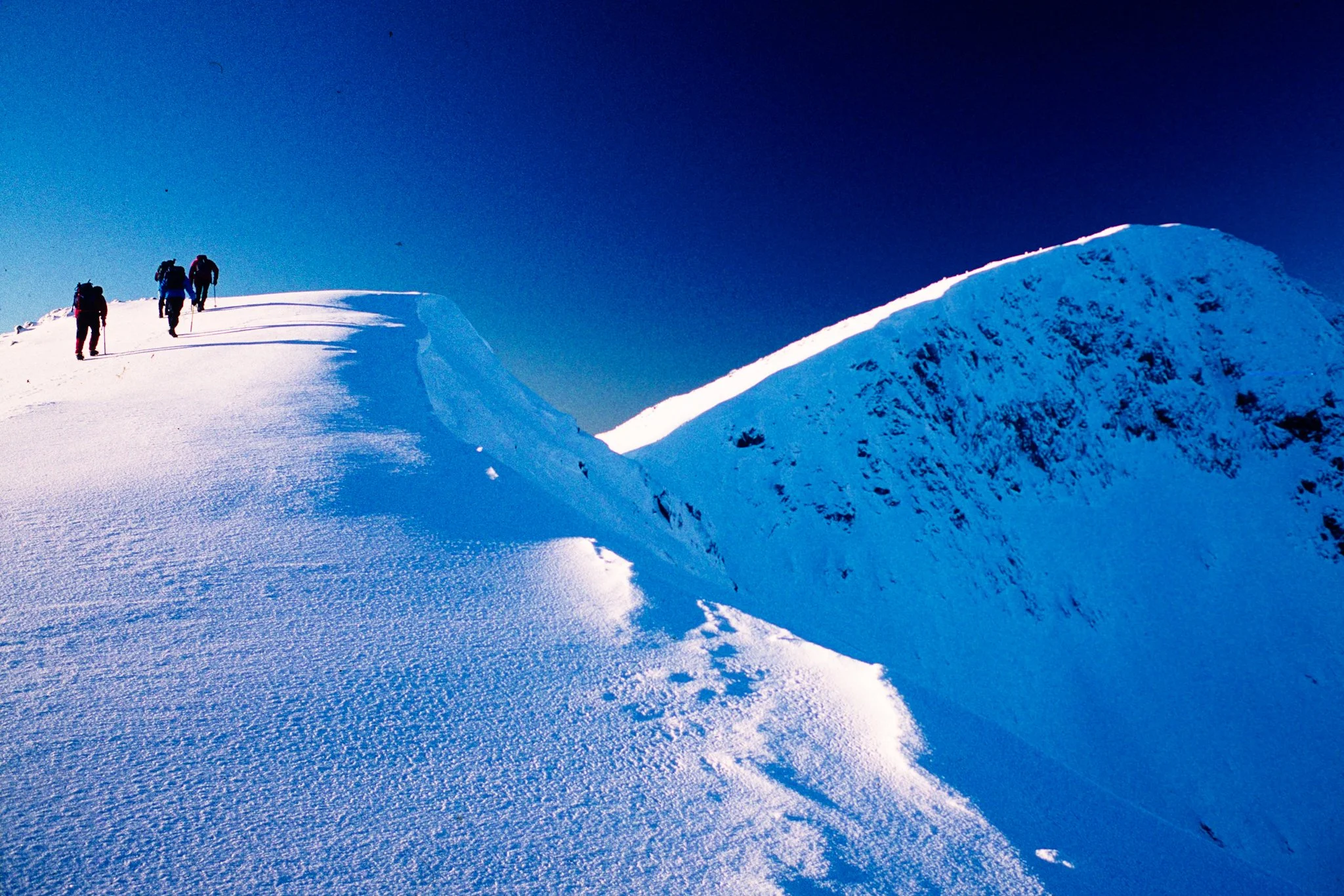 Four climbers ascending a snow-covered mountain ridge against a clear blue sky.
