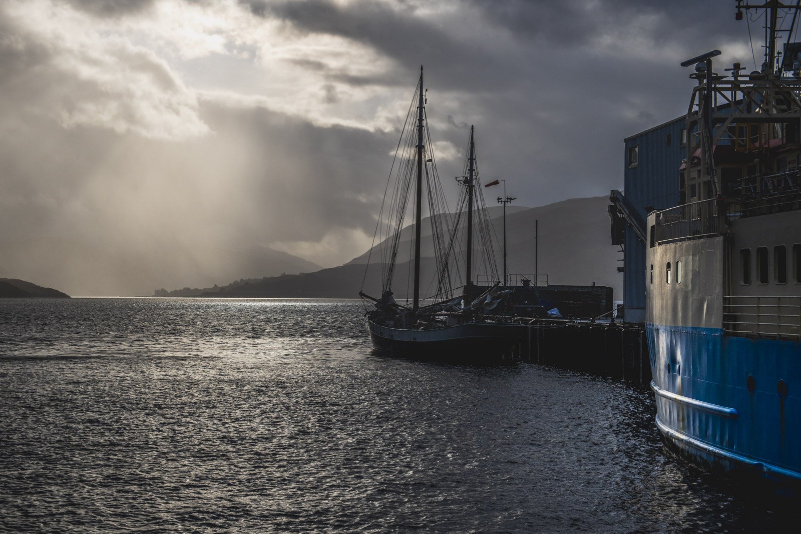 A docked ship and sailboat on a waterway during a cloudy day with sunlight breaking through the clouds and distant mountains in the background.