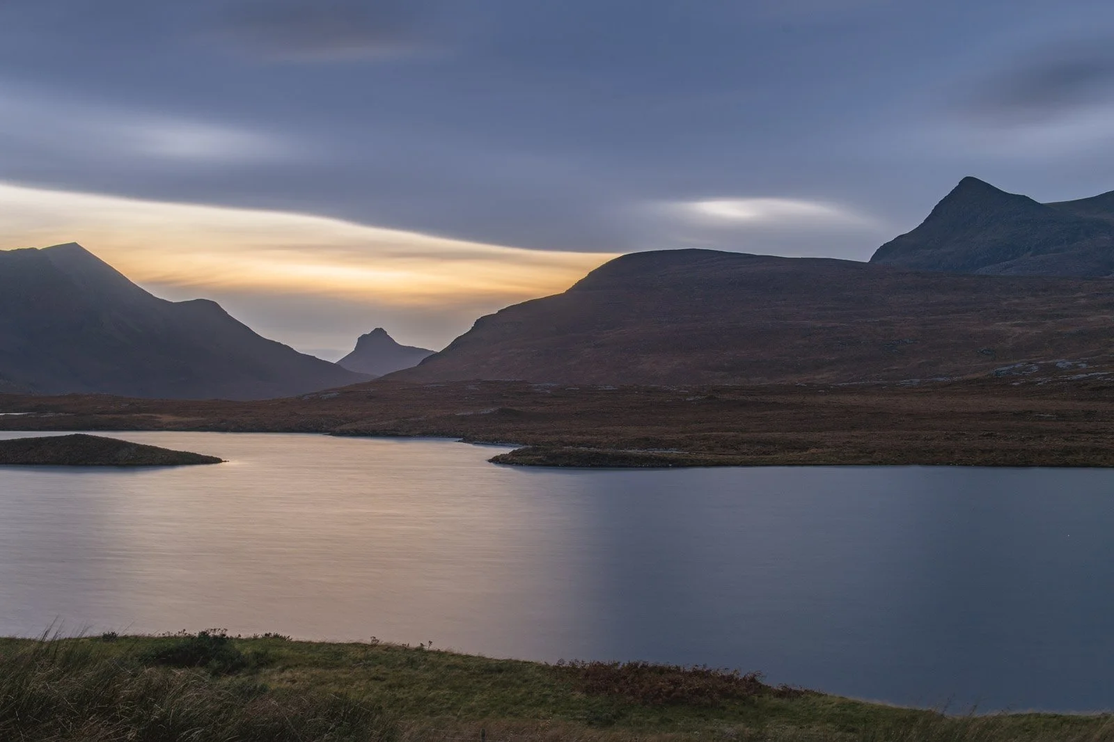 A scenic landscape of mountains and a lake at sunset or sunrise, with dark clouds overhead and a glow on the horizon.