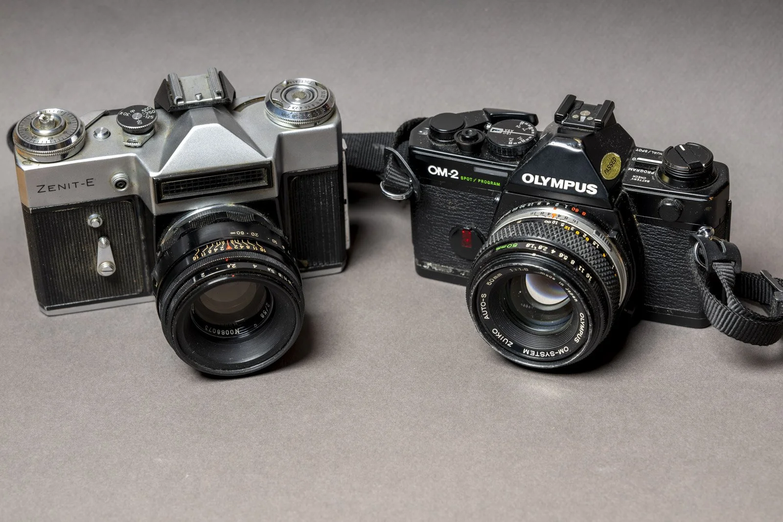 Two vintage film cameras on a gray surface. The camera on the left is a silver Zenit-E, and the one on the right is a black Olympus OM-2 Spot Program