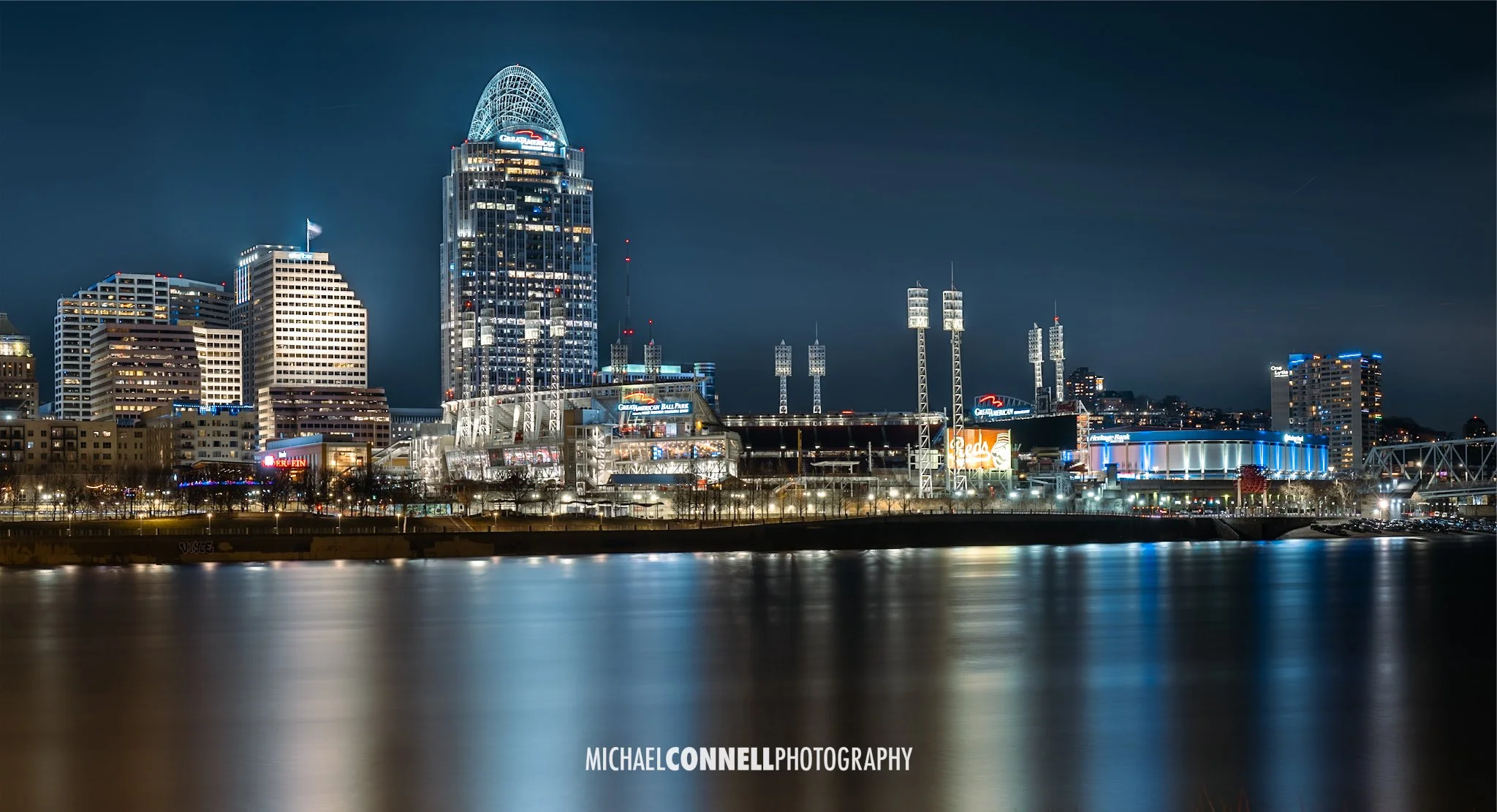 Nighttime cityscape of Cincinnati with illuminated skyscrapers, including the Great American Tower at Queen City Square, reflecting on the Ohio River.