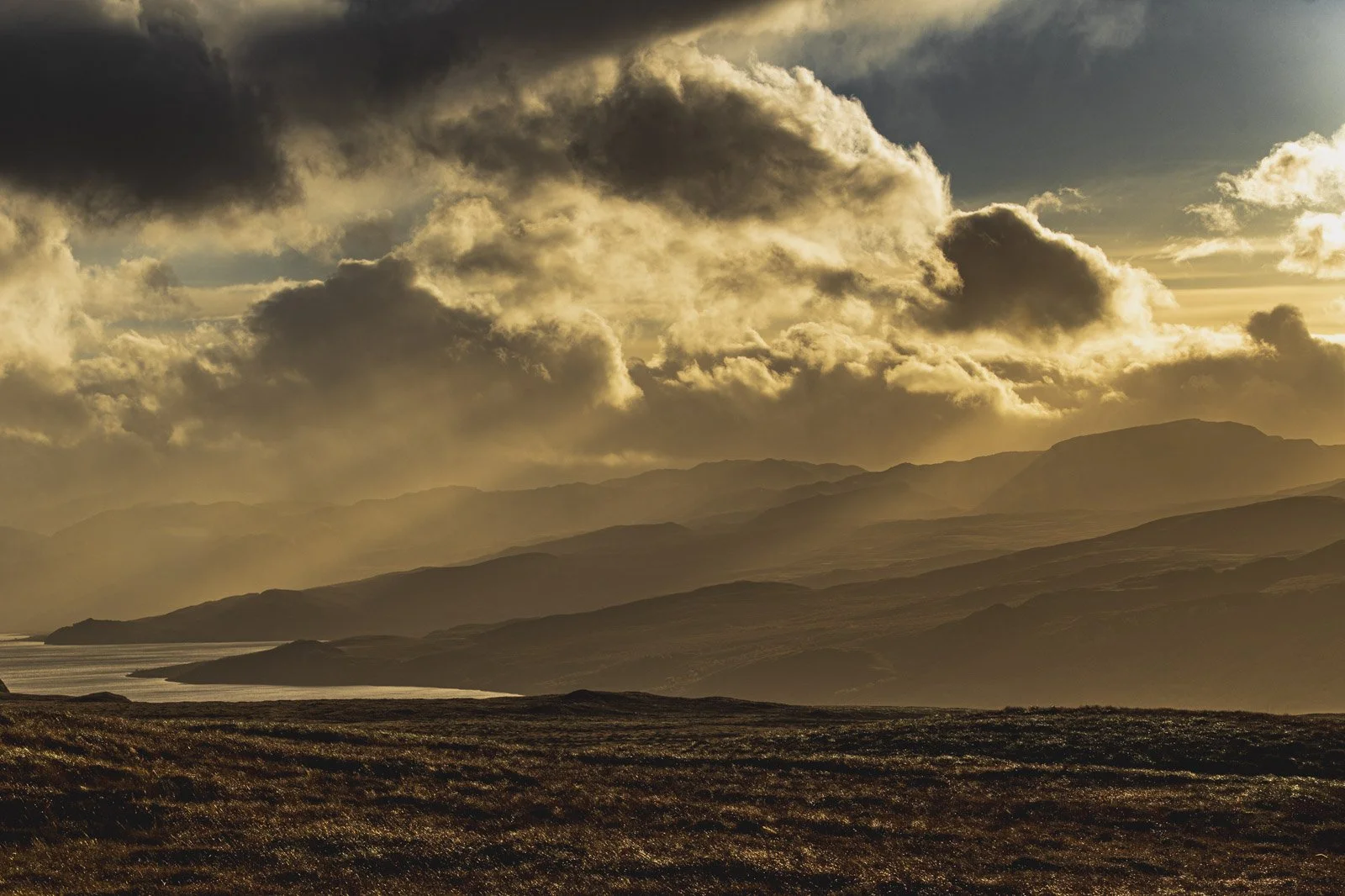 A landscape of rolling hills and mountains under a dramatic sky with dark and light clouds during sunset or sunrise.