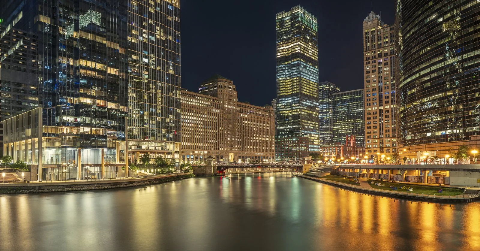 Night view of skyscrapers reflecting on a river in a city, with illuminated windows and lights.